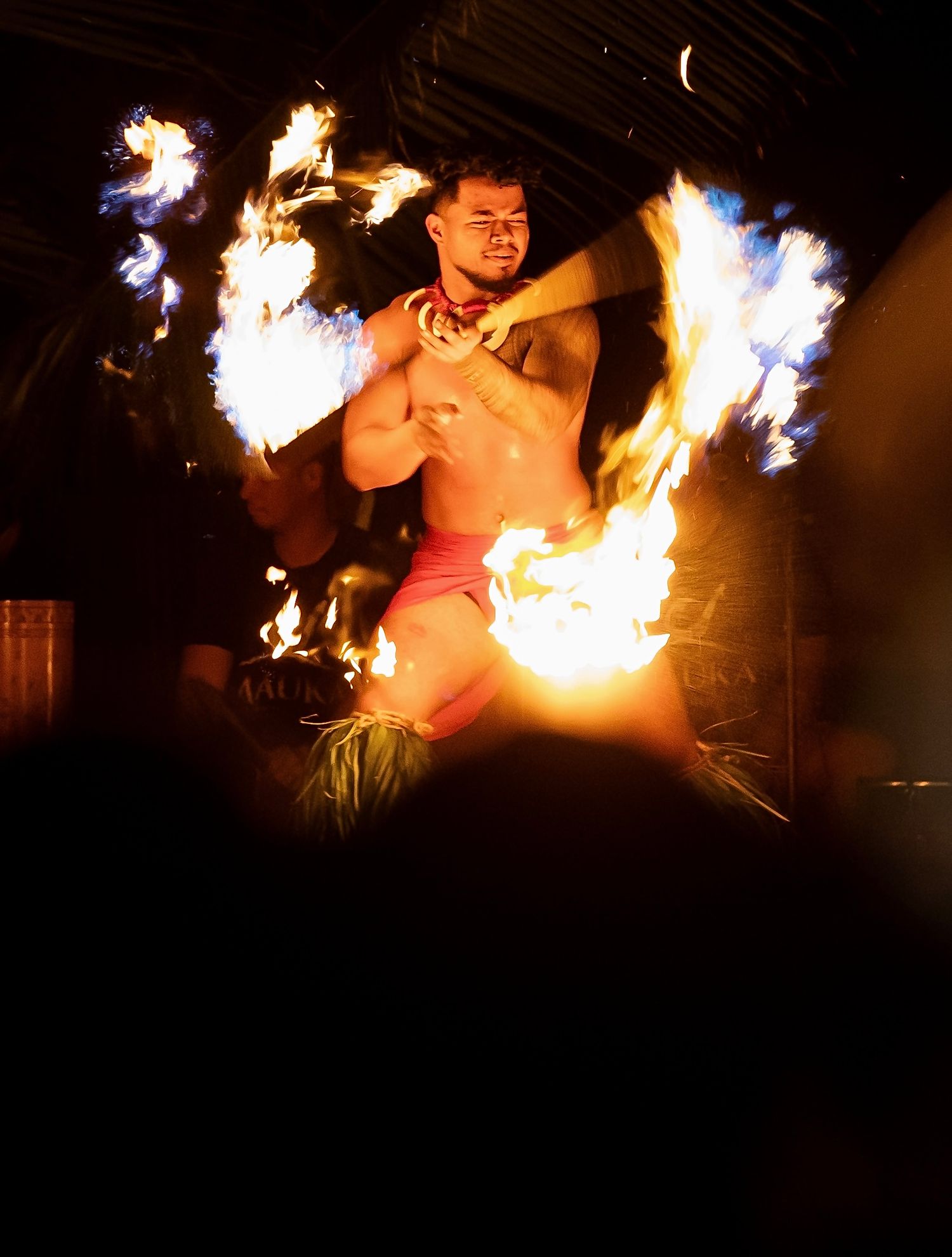 A shirtless fire dancer performs a traditional Polynesian fire knife dance, wielding flaming batons before an audience.