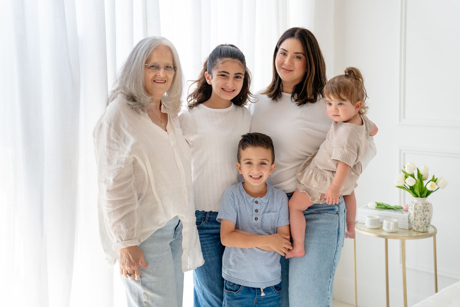 Multi-generational family of five smiling together in a bright white room, including grandmother, mother, and three children.
