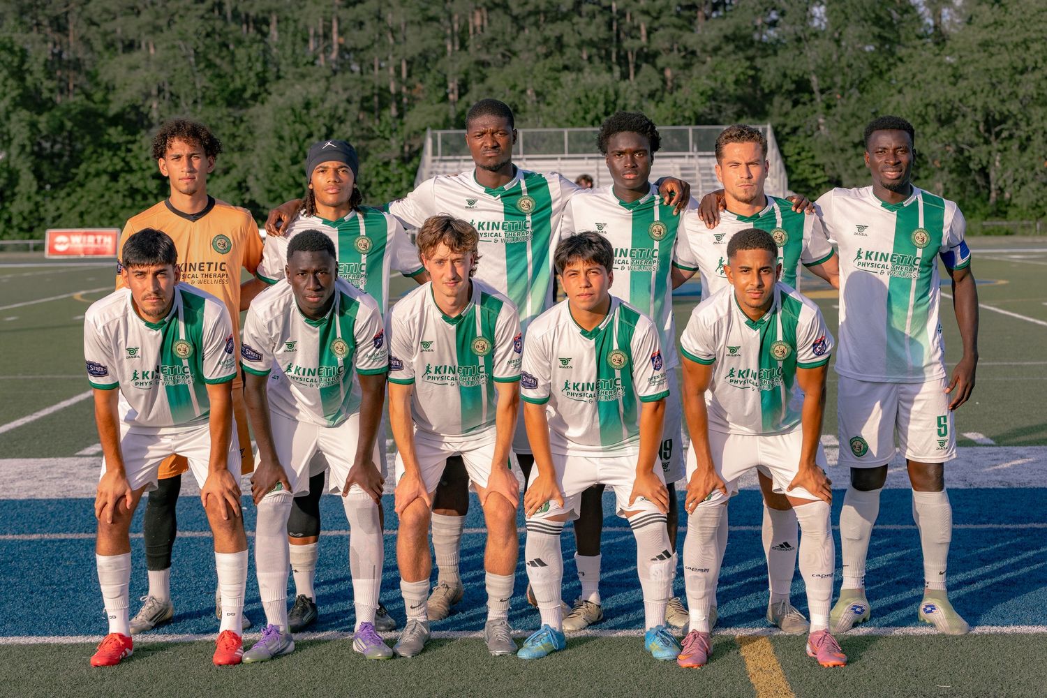 Youth soccer team poses for group photo in white and green uniforms on outdoor field at dusk.