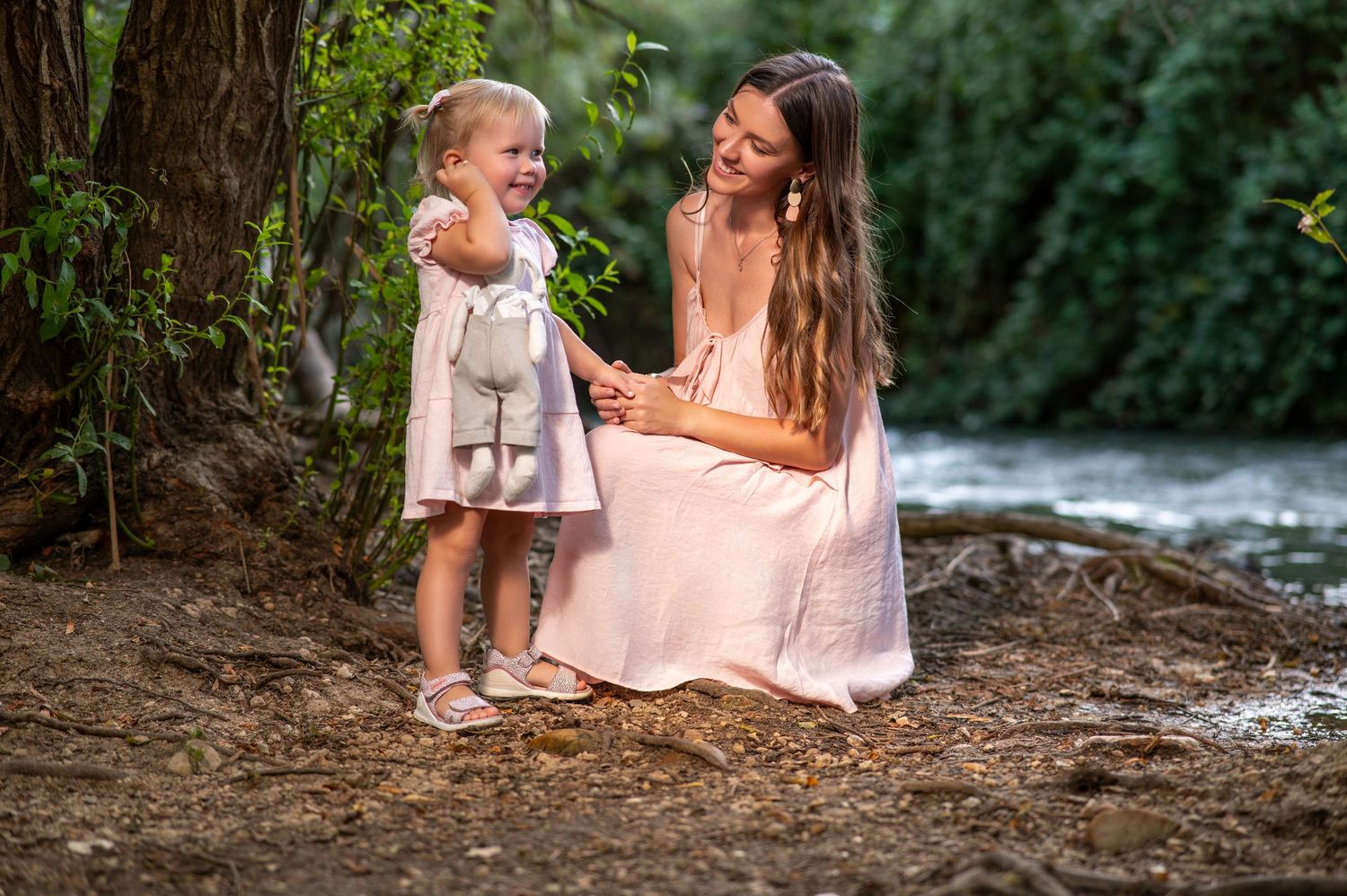 A tender moment shared between a woman in a pink dress and a small child by a peaceful stream in a forest setting.