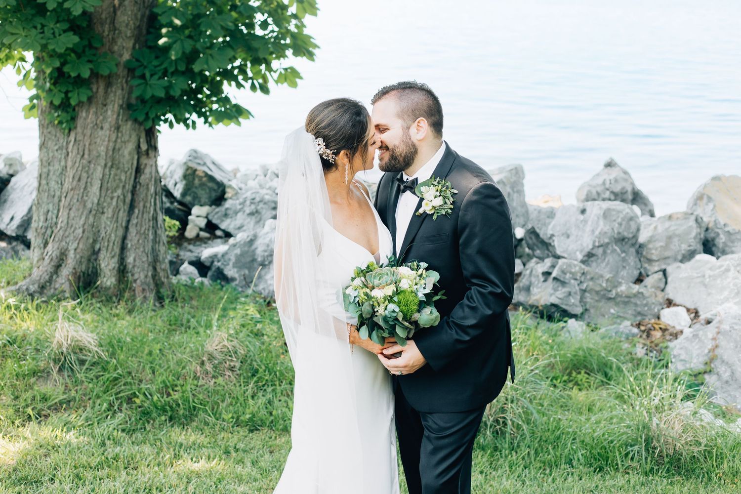 A romantic waterfront wedding portrait with a couple embracing near rocky shoreline under a large tree.