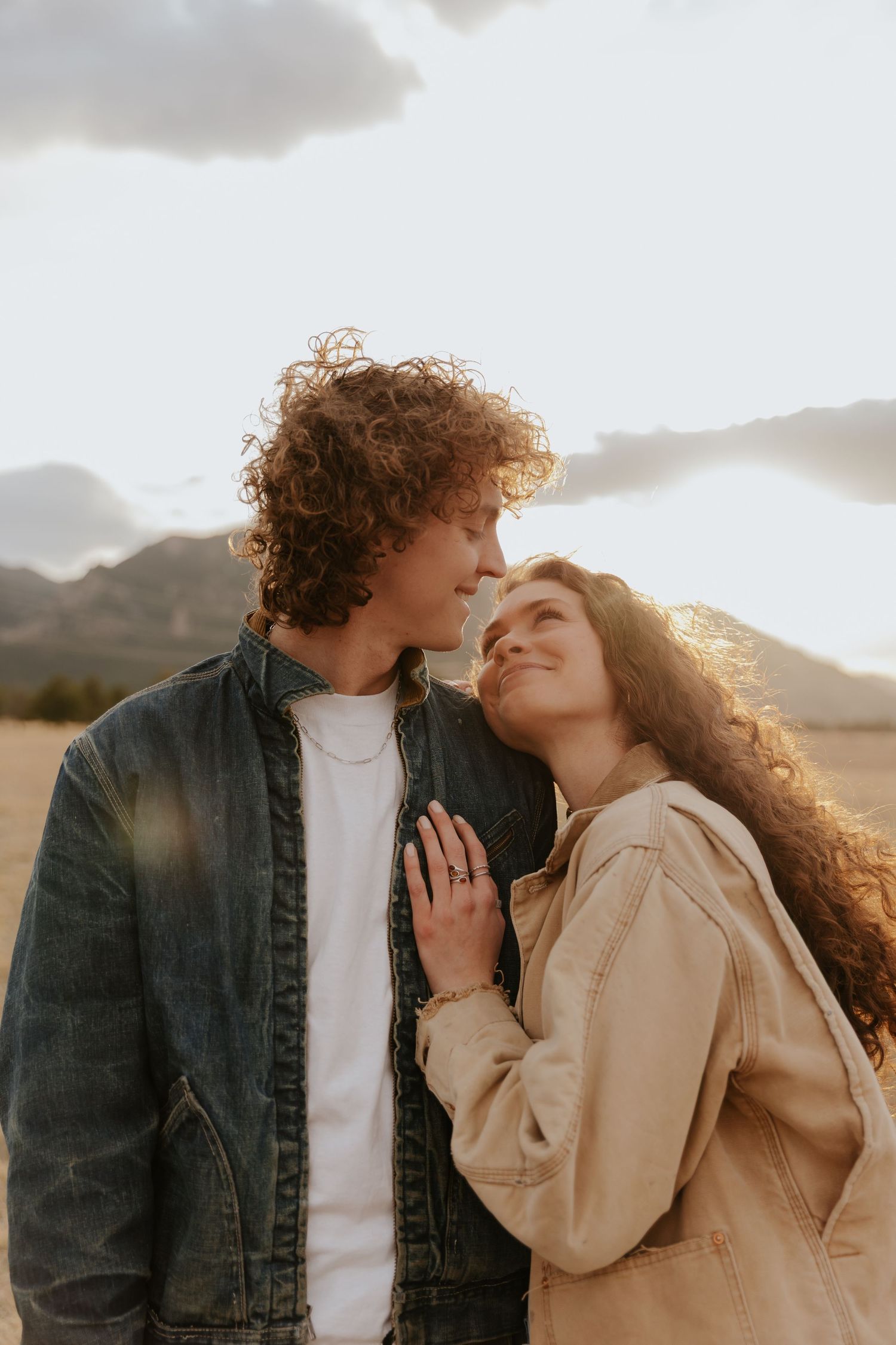 Two people in casual jackets sharing an intimate moment outdoors against a mountain backdrop at sunset.