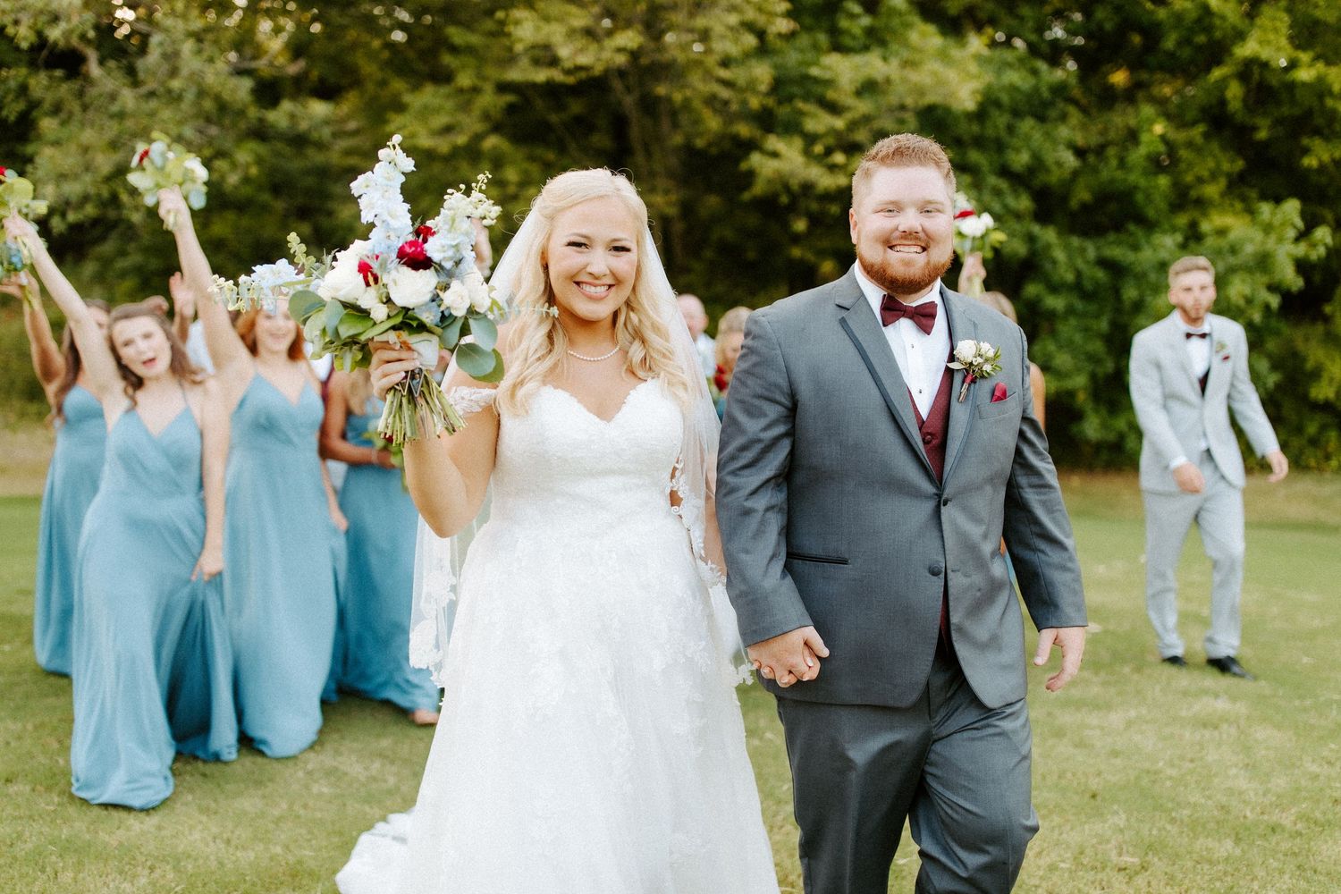 A joyful wedding party celebrates on a green lawn, with bridesmaids in light blue dresses following the newlyweds.