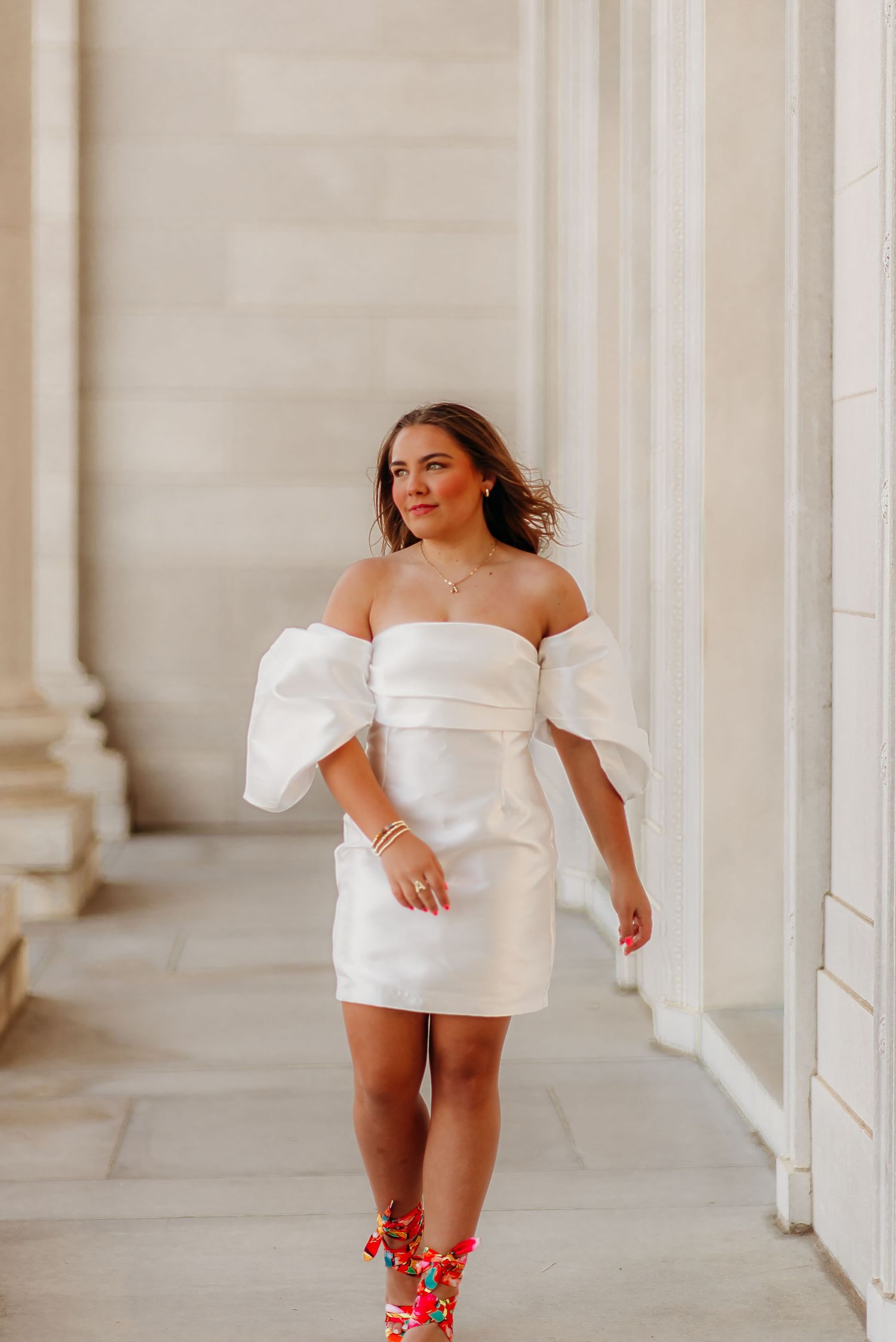 A person in a white off-shoulder mini dress walks along a colonnade with red floral boots.