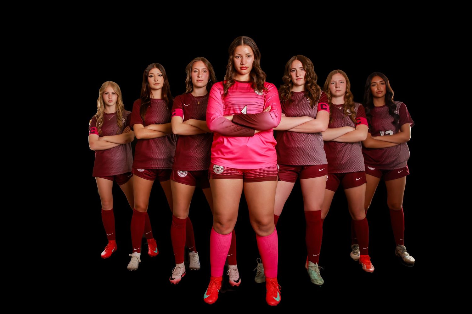 A group of soccer players in red uniforms pose together against a black background for a team photo.