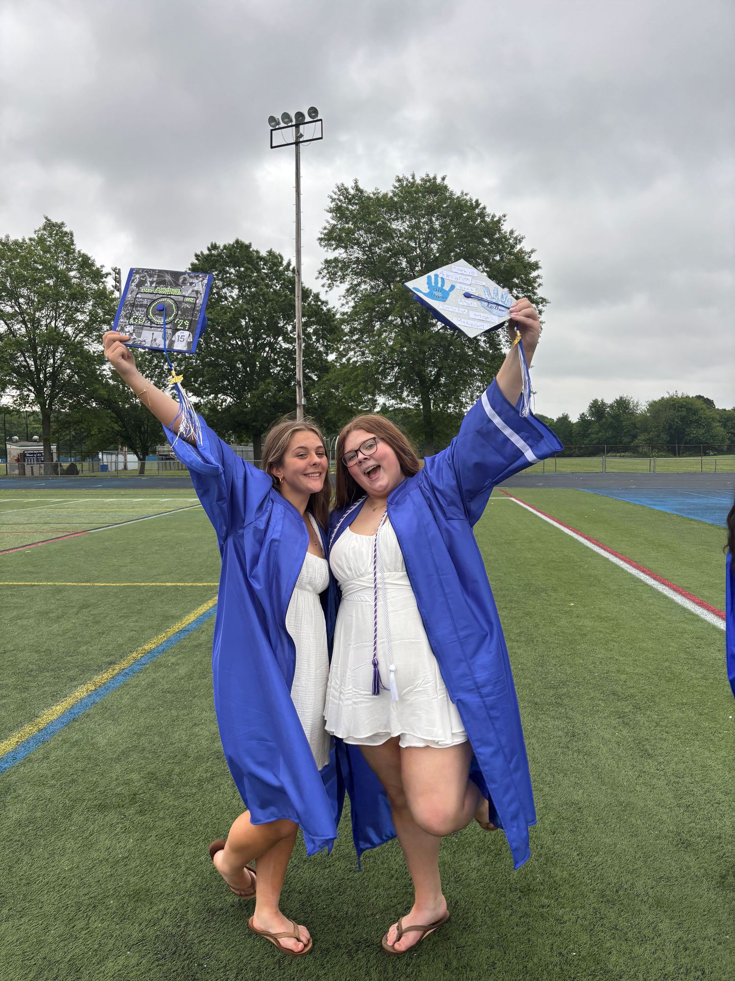 Two graduates celebrate in blue robes on sports field.