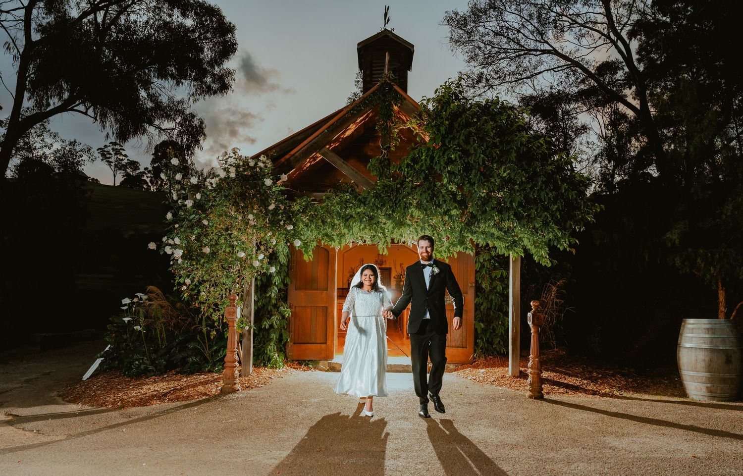 Two people walk together down a pathway in front of a rustic wooden chapel illuminated at dusk.