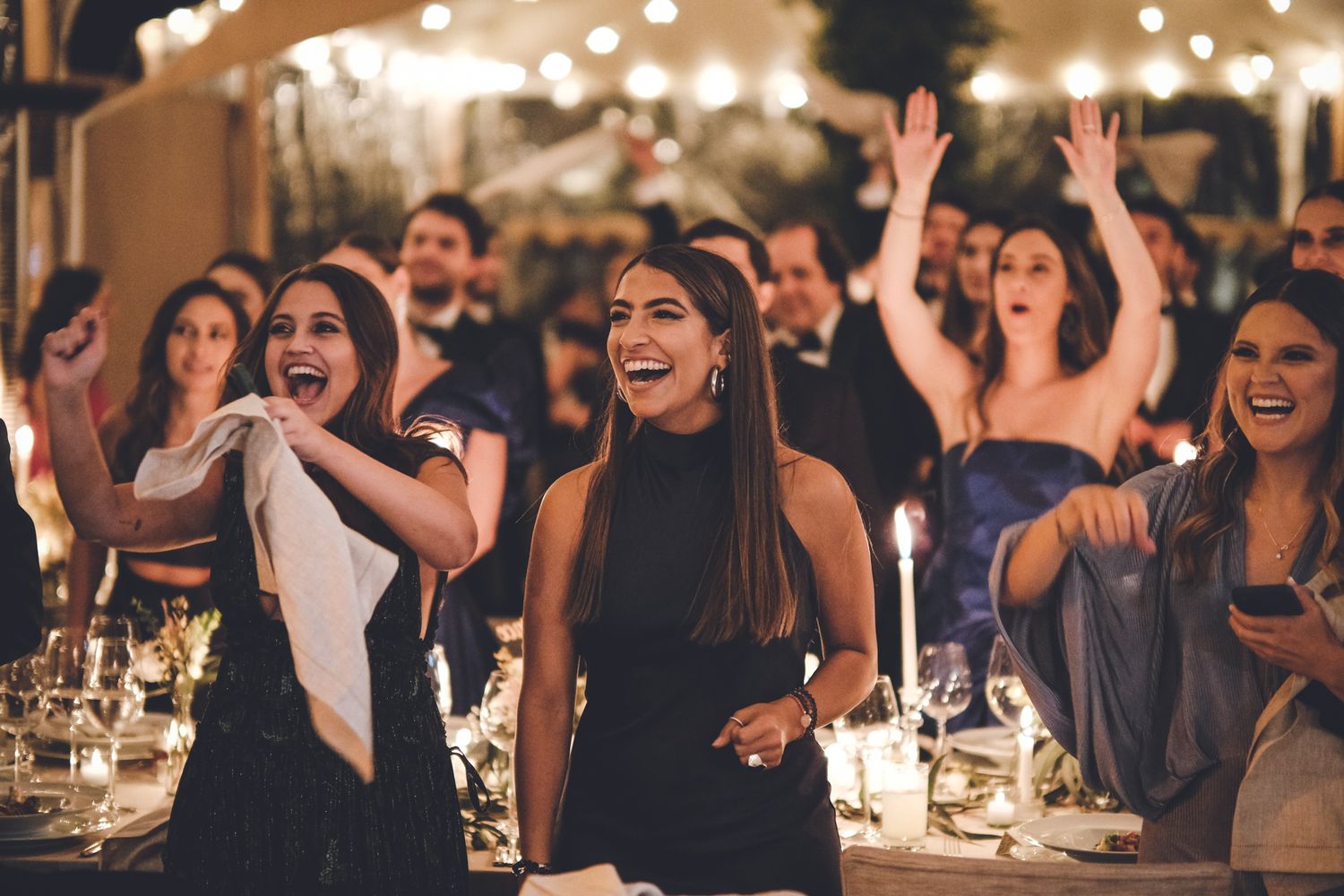 A group of party guests celebrate and dance enthusiastically at an evening reception with candlelit tables.