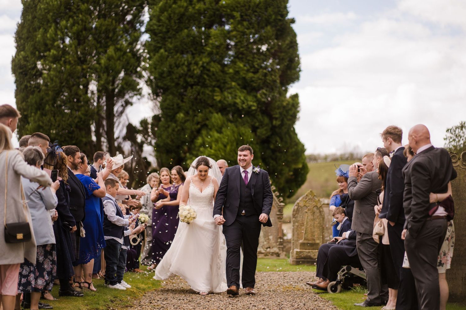 Wedding party and guests line a countryside path amidst tall cypress trees during an outdoor celebration.