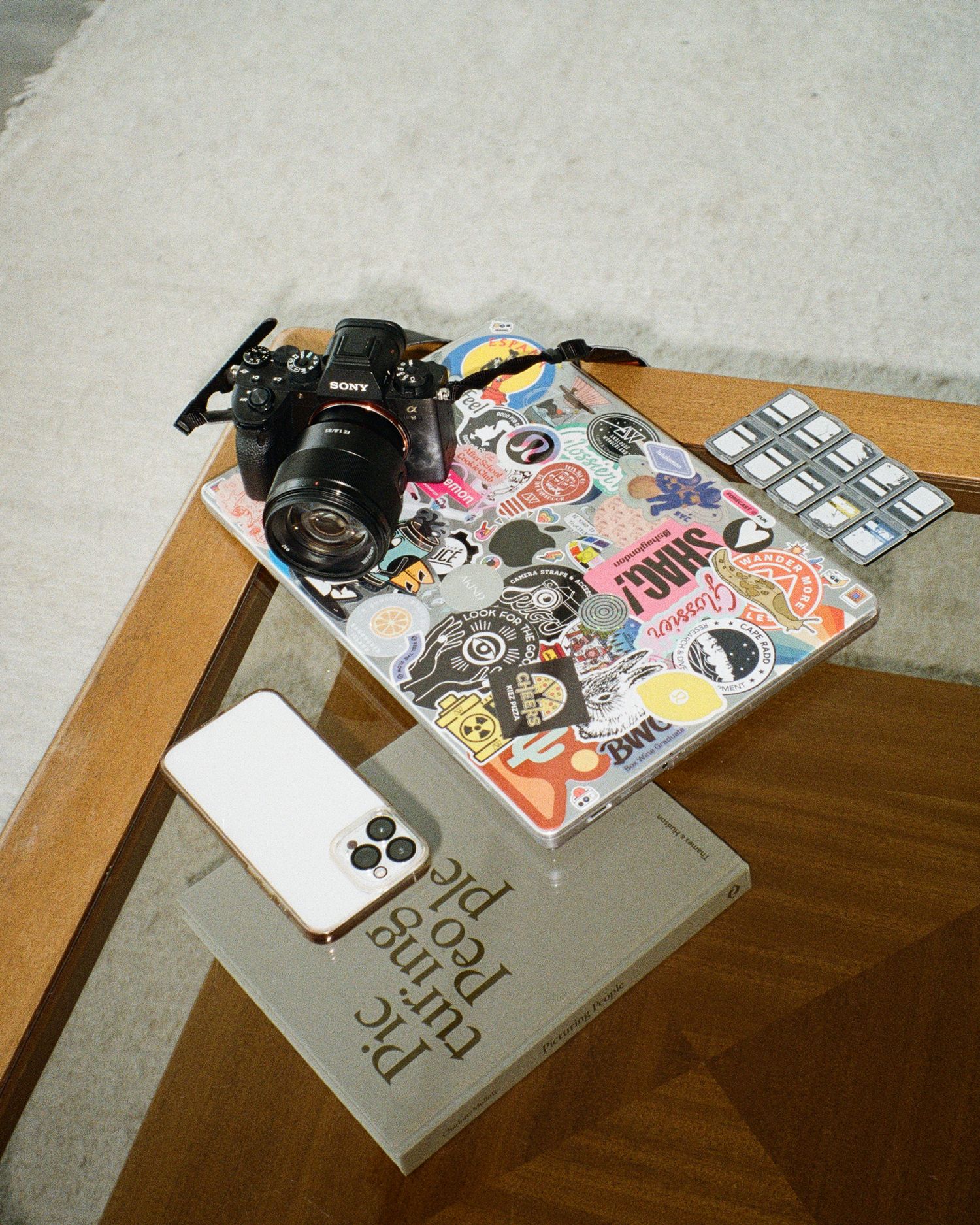 A Canon camera sits next to a laptop covered in stickers and a white iPhone on a wooden table.
