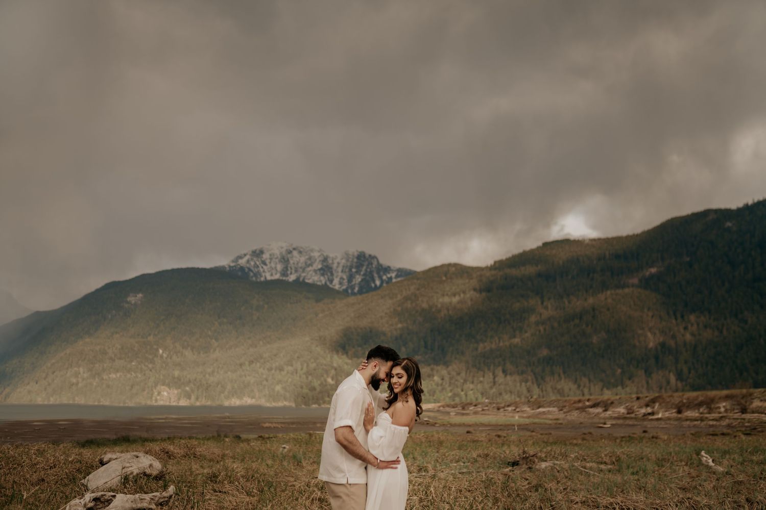 A couple shares an intimate moment on a rocky shore with dramatic mountains and moody clouds in the background.