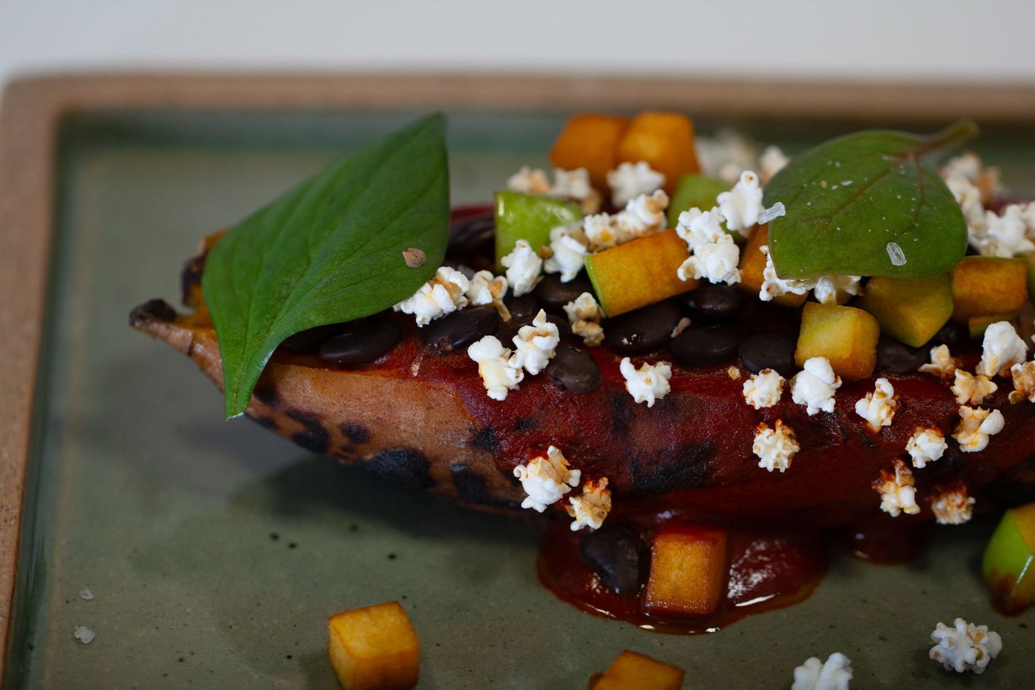 Roasted sweet potato garnished with mini popcorn, green apple cubes and fresh basil leaves on a green ceramic plate.