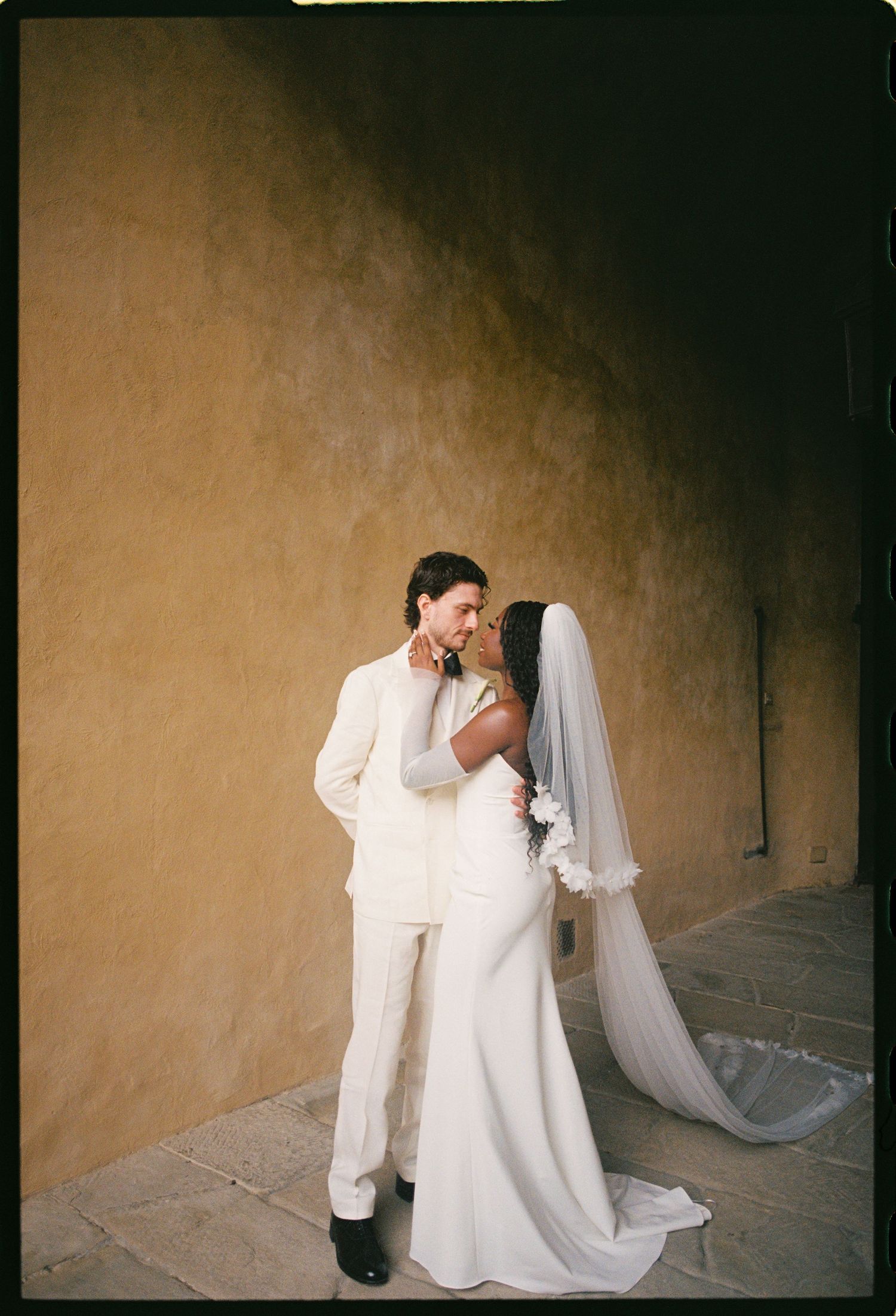 A couple in matching white attire share an intimate moment during their wedding against a rustic beige wall.