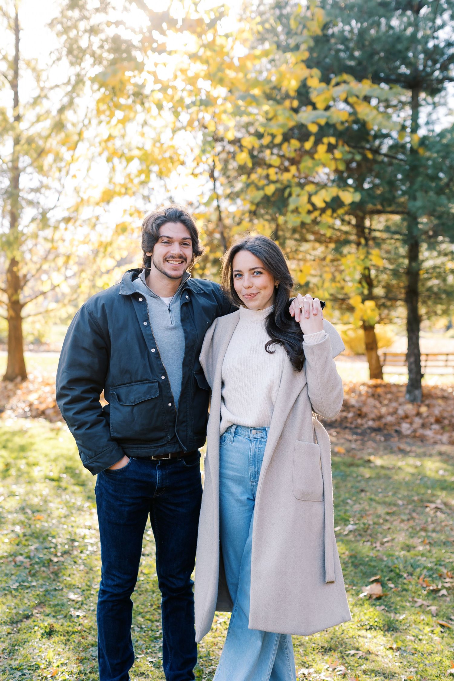 A couple stands together on an autumn day surrounded by trees with golden leaves.