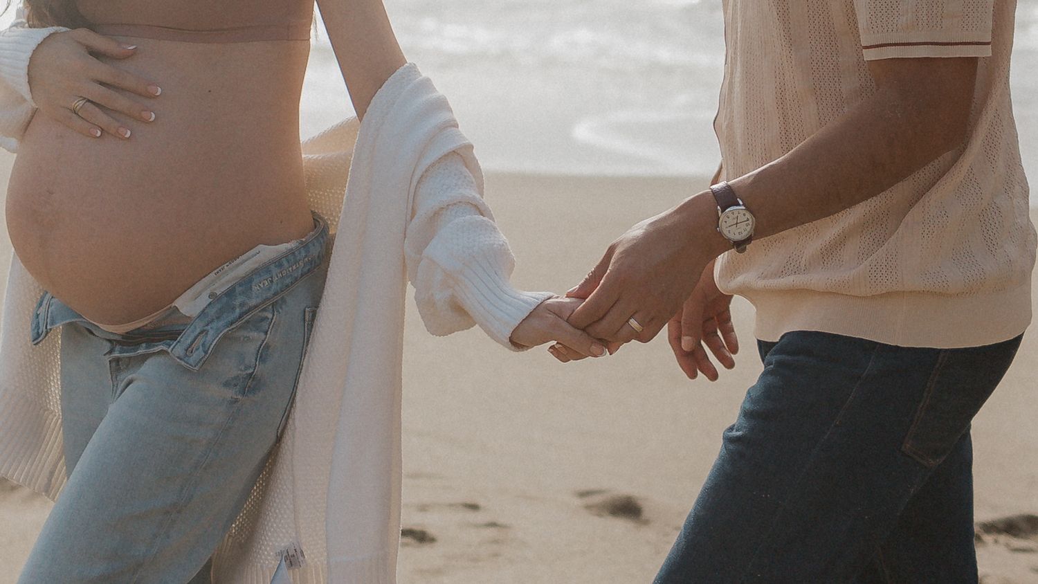 Close-up of expecting couple holding hands on beach at sunset.