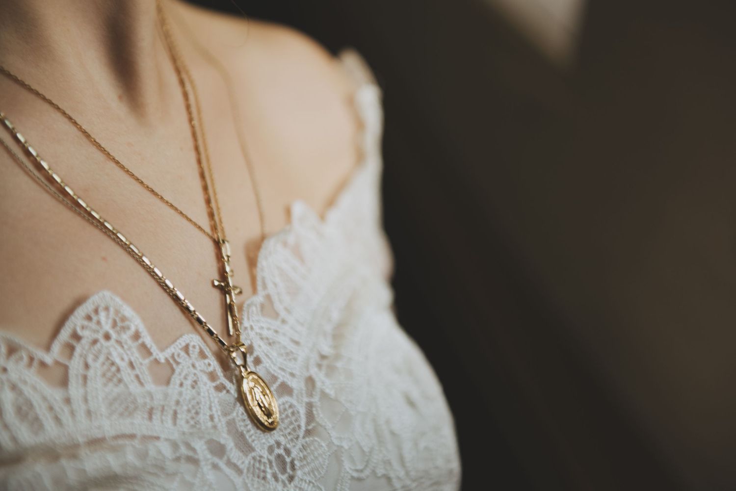 a bride wears a gold crucifix and miraculous medal around her neck while wearing a lace bridal gown.