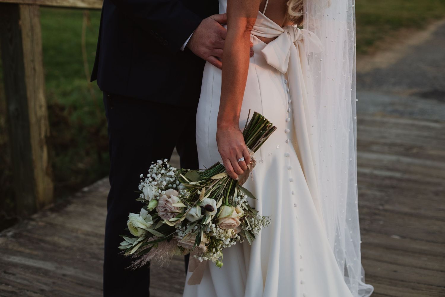 Close-up view of elegant white wedding bouquet held against a flowing bridal gown on wooden walkway.