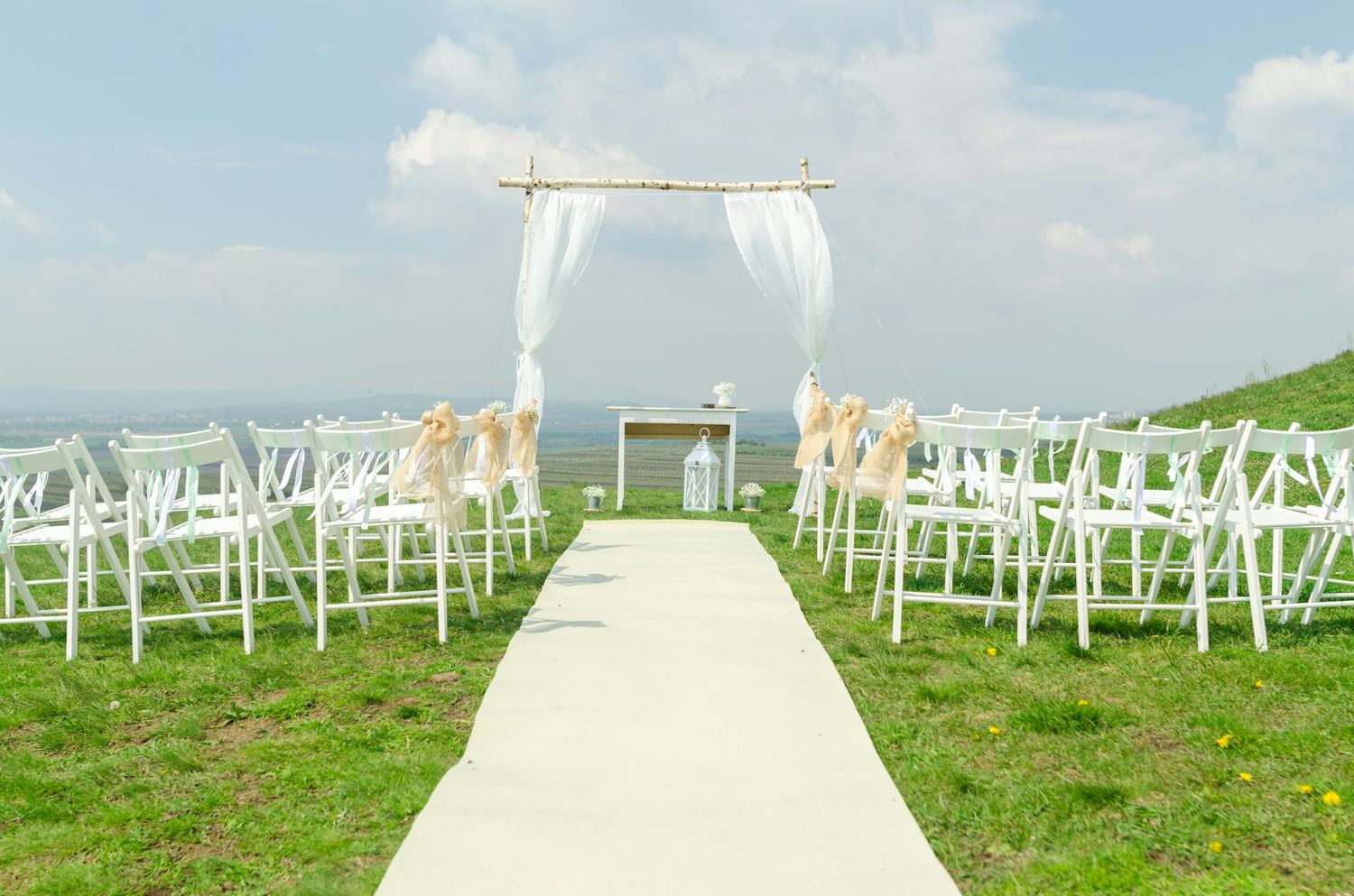 White fabric arch and rows of white chairs set up for an outdoor wedding ceremony on a grassy hillside by the ocean.