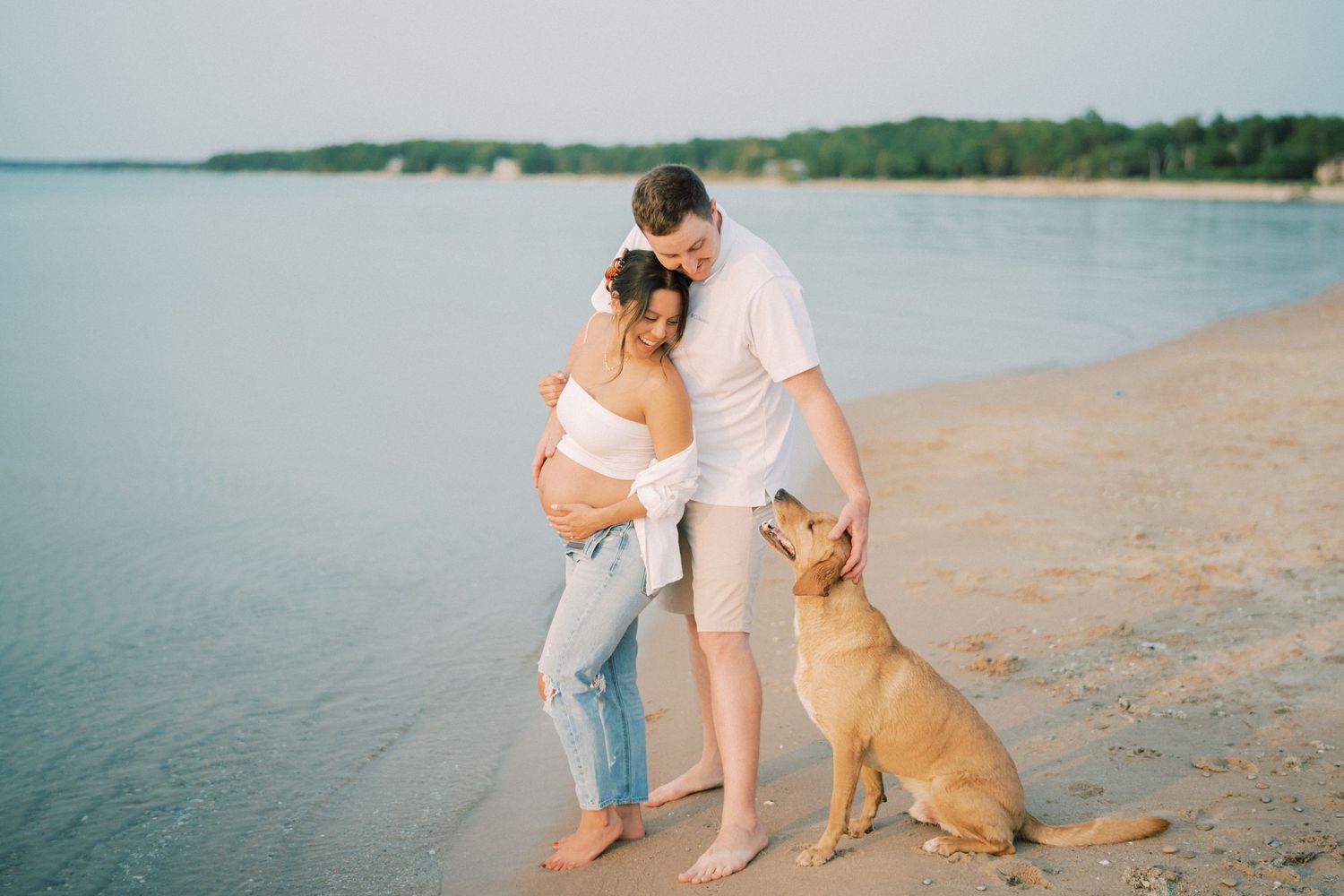 A couple embraces on the beach with their golden retriever as the sun sets over the water.