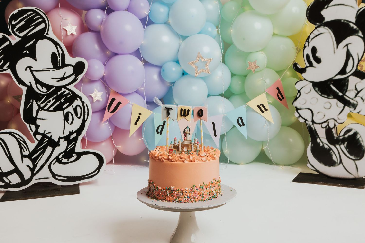 Mickey Mouse themed birthday cake with rainbow balloon arch and black and white character cutouts.