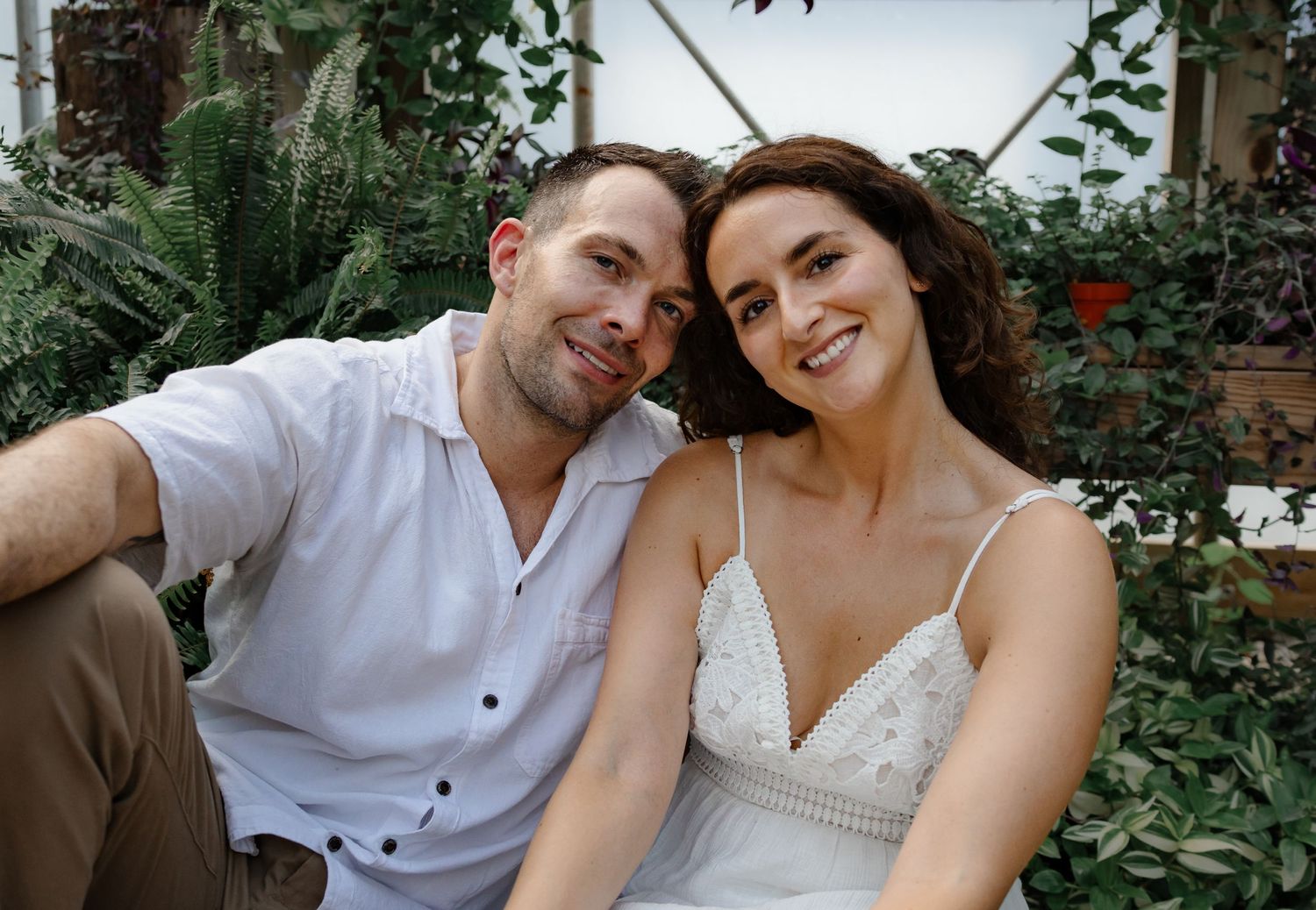 A couple sits together in a garden setting with lush greenery in the background.