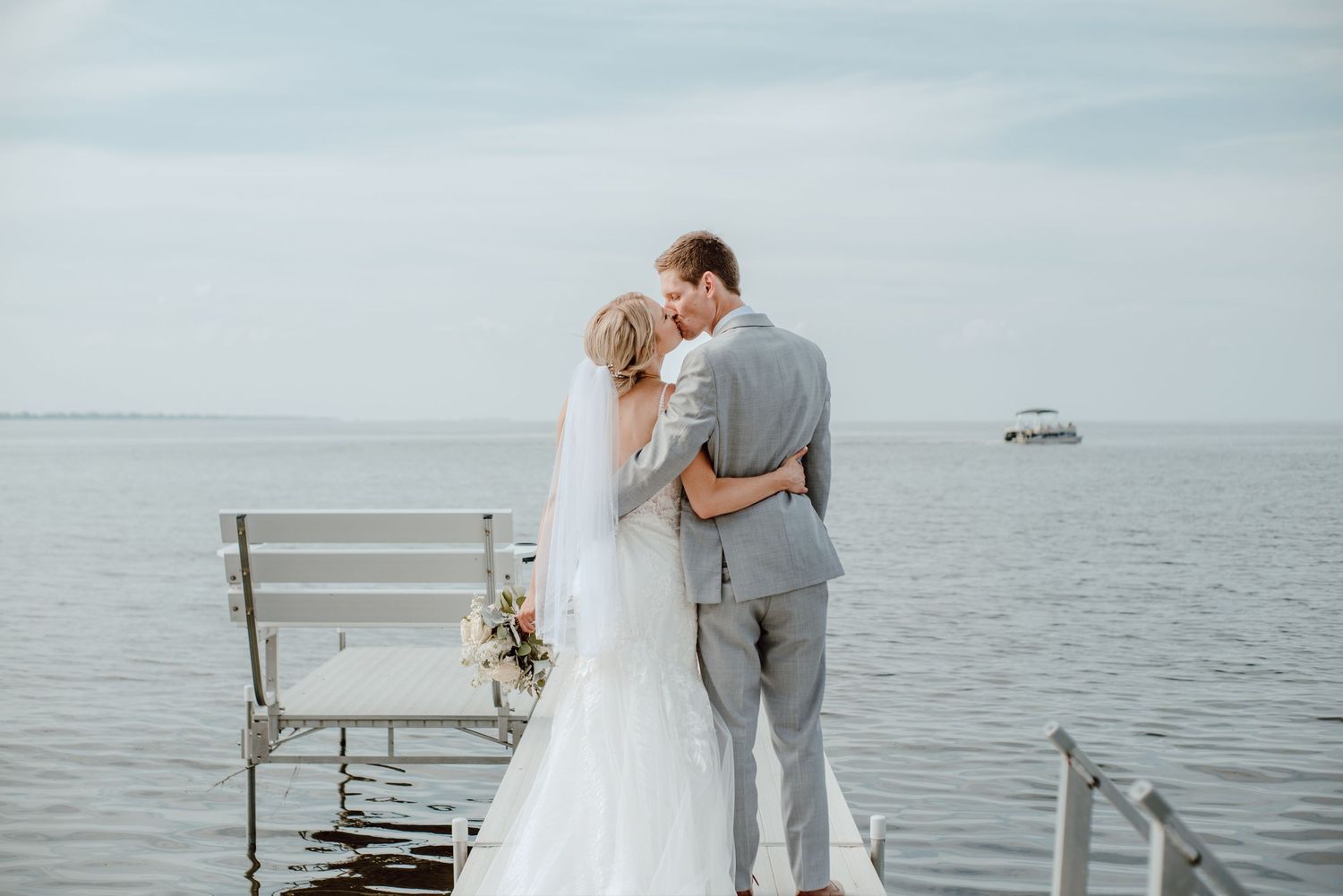A romantic wedding couple embraces on a dock overlooking calm waters with a boat visible in the distance.