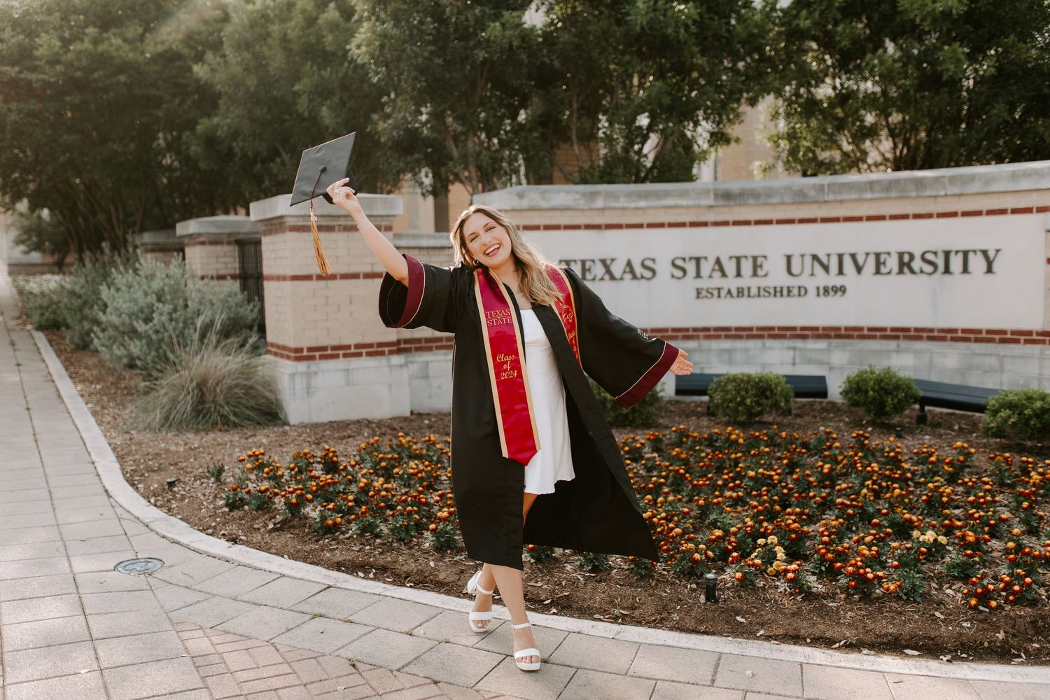 Graduate in cap and gown celebrates joyfully in front of Texas State University sign with orange flowers below.