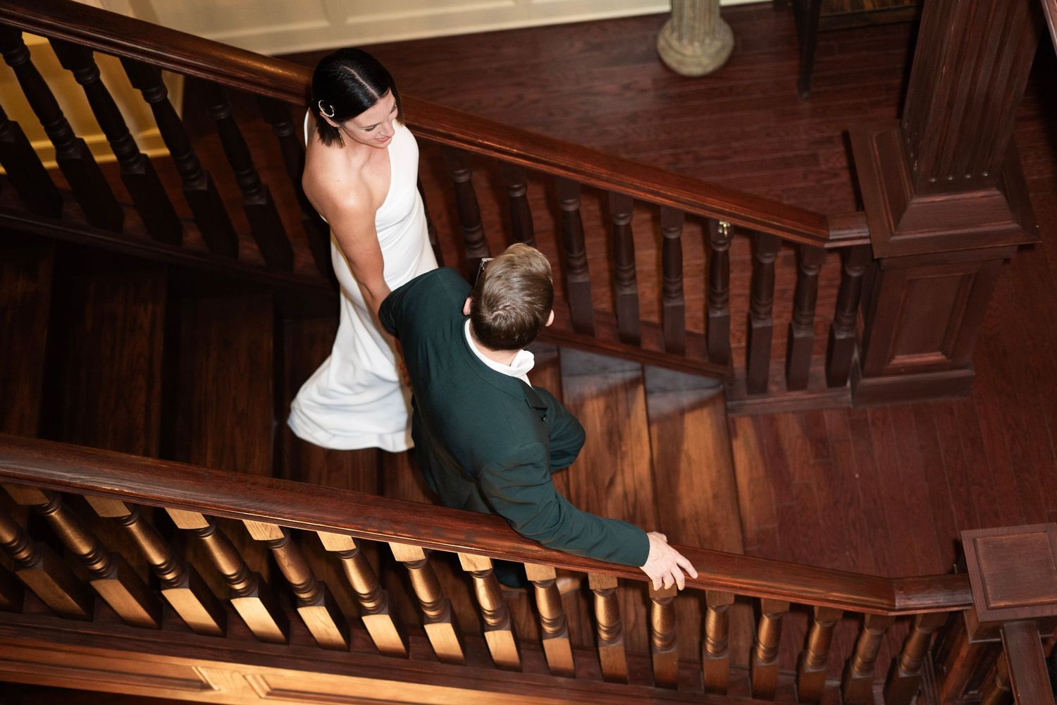 Aerial view of bride in white gown and groom in green suit holding hands on elegant wooden staircase.