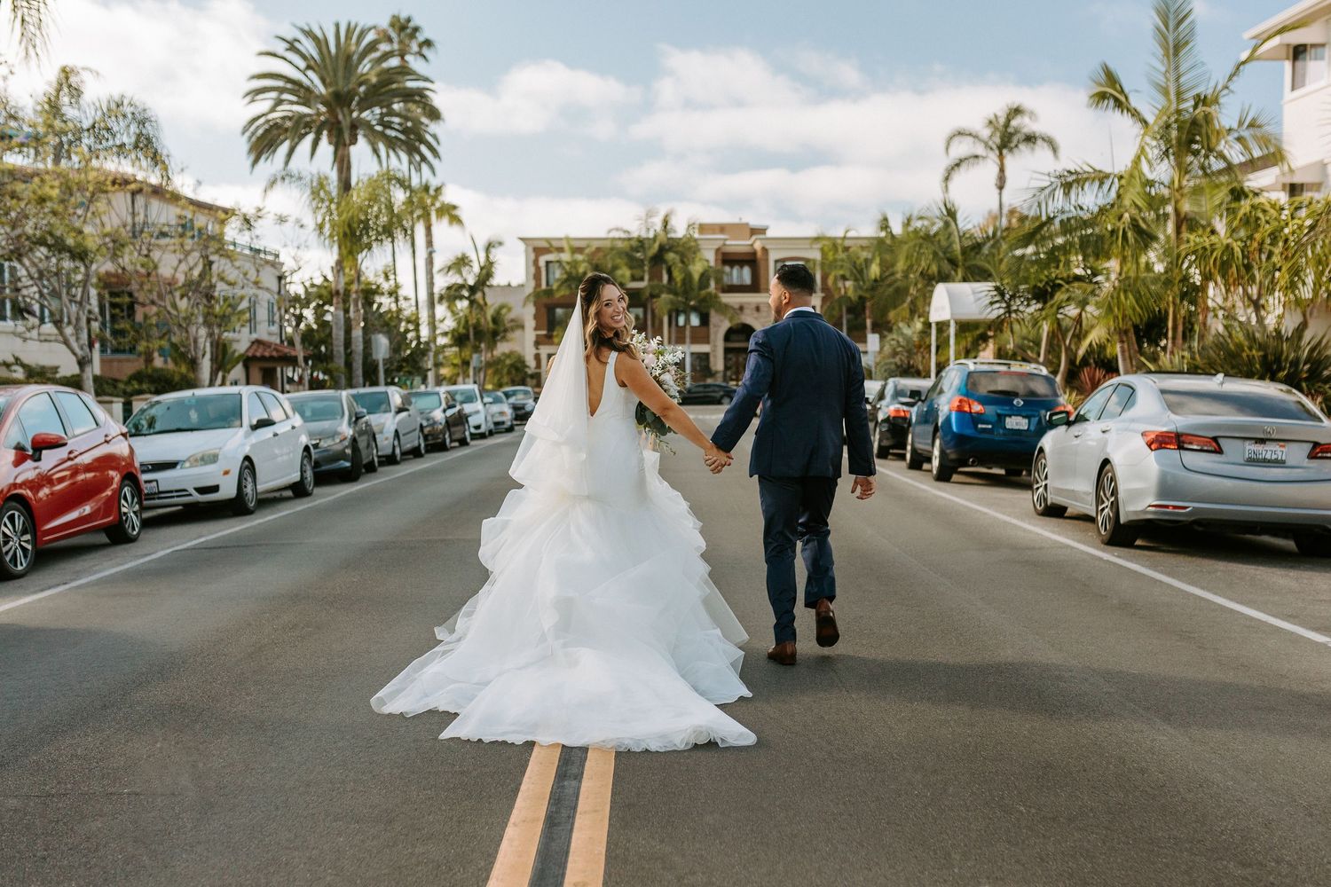 Bride and groom walk hand-in-hand down a palm-lined street in wedding attire, with parked cars and buildings in the background in La Jolla.