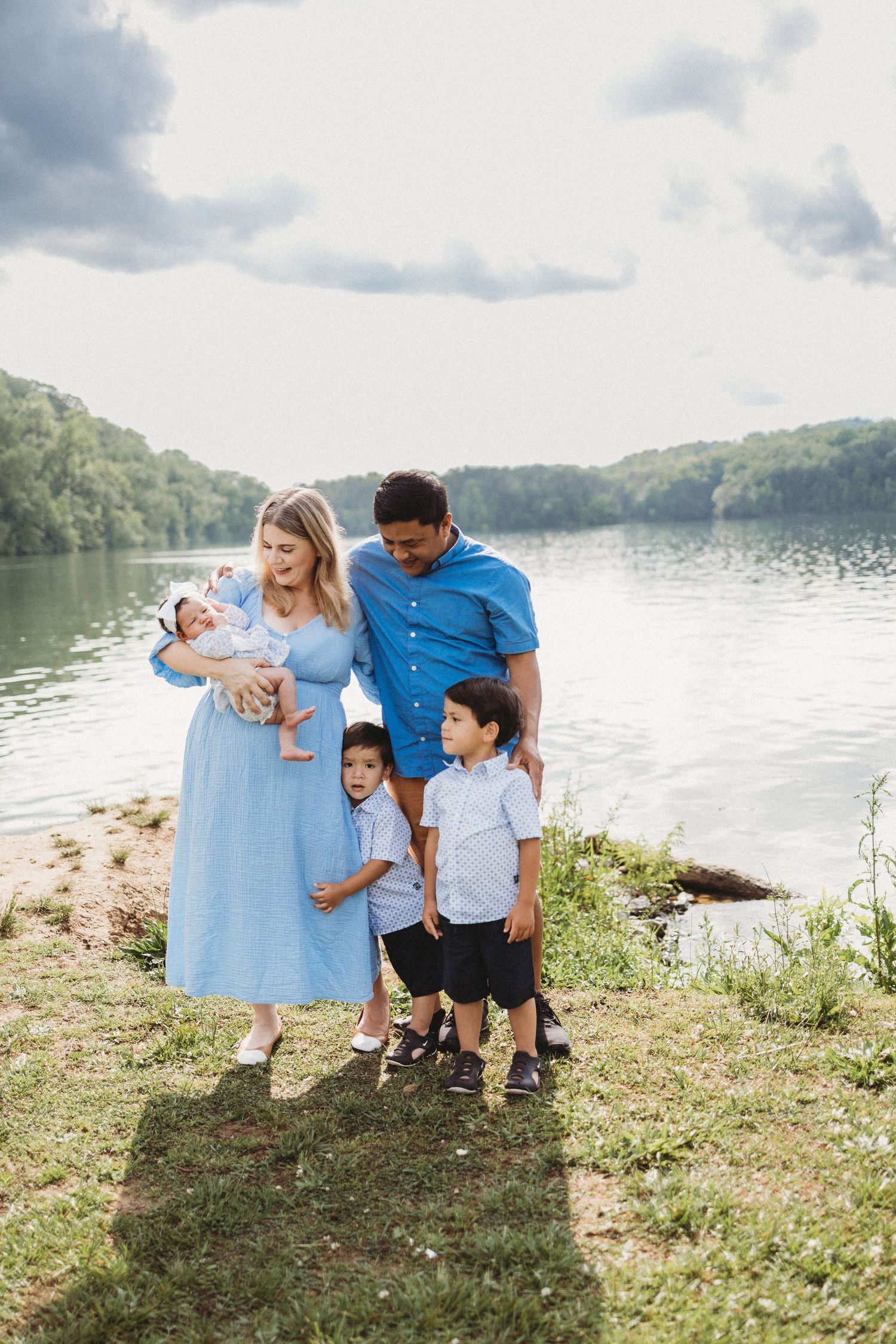 Family of five posing by a serene lake, mother in blue dress holding newborn, father and two young boys beside her.