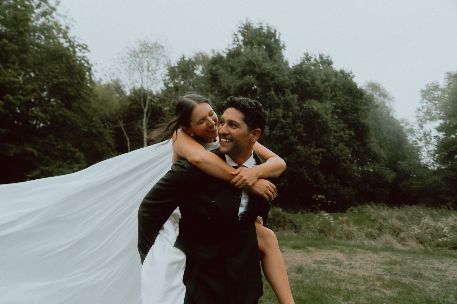 A joyful groom carries his bride piggyback in a lush green field, her white veil flowing in the wind.