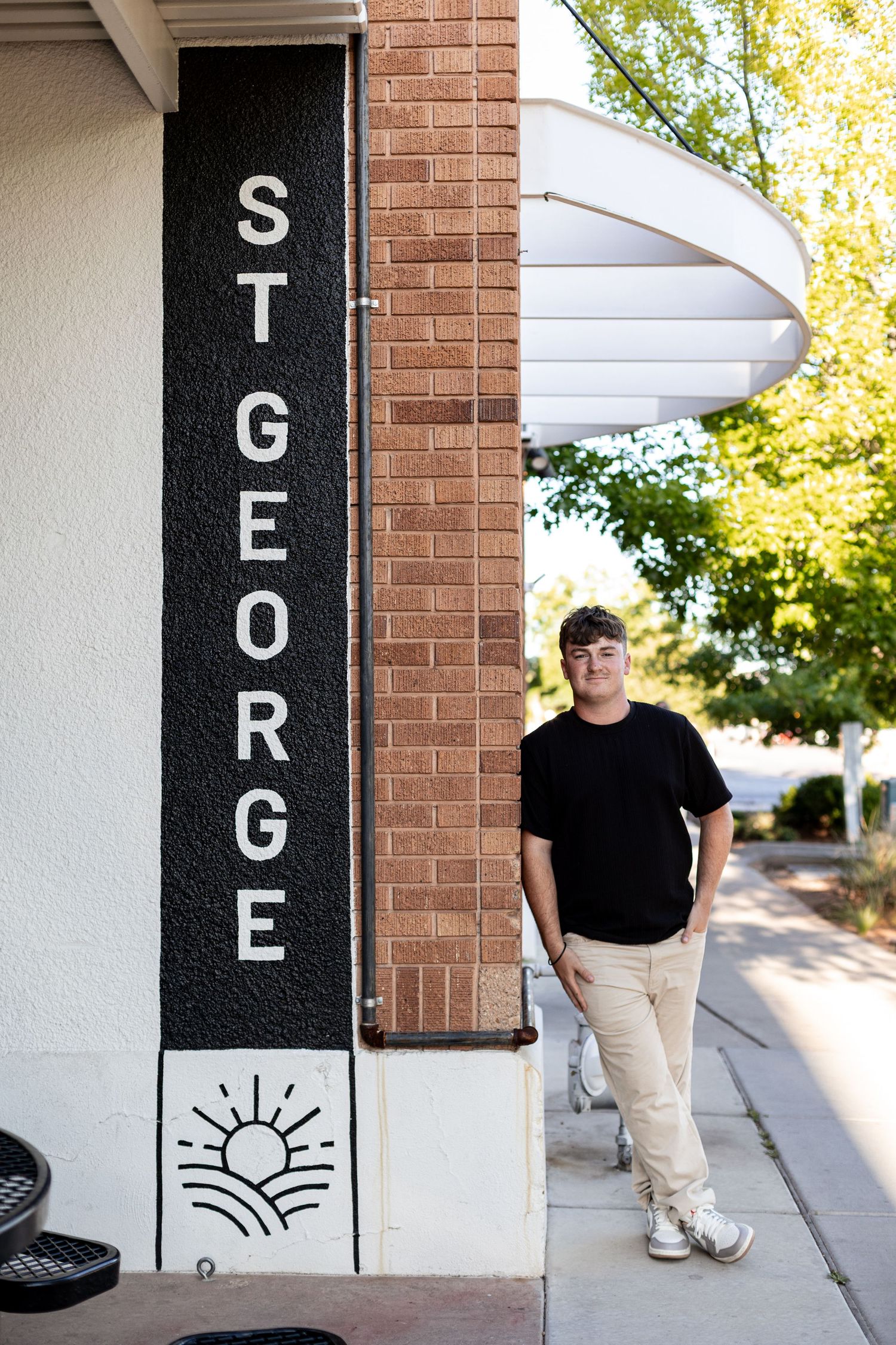 A young man in a black t-shirt stands beside a vertical St. George sign on a brick wall along a sunny sidewalk.