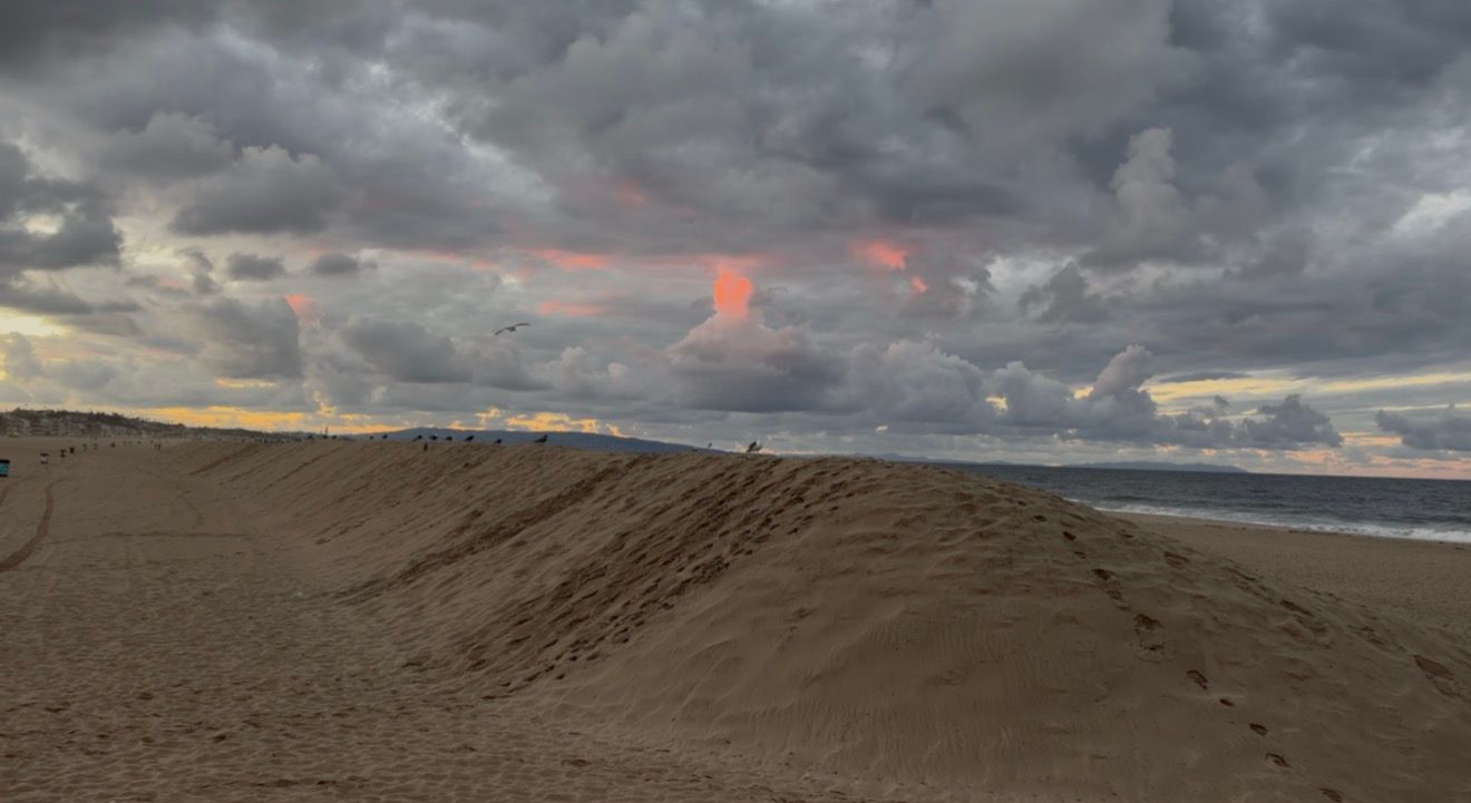 Sandy beach dunes at sunset under a dramatic cloudy sky with pink and orange glowing clouds above the calm ocean horizon.