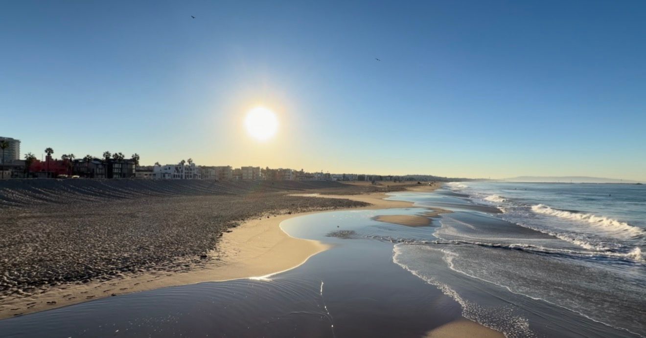 Bright sun shines over a quiet sandy beach with gentle waves rolling ashore and coastal buildings lining the distant shoreline.