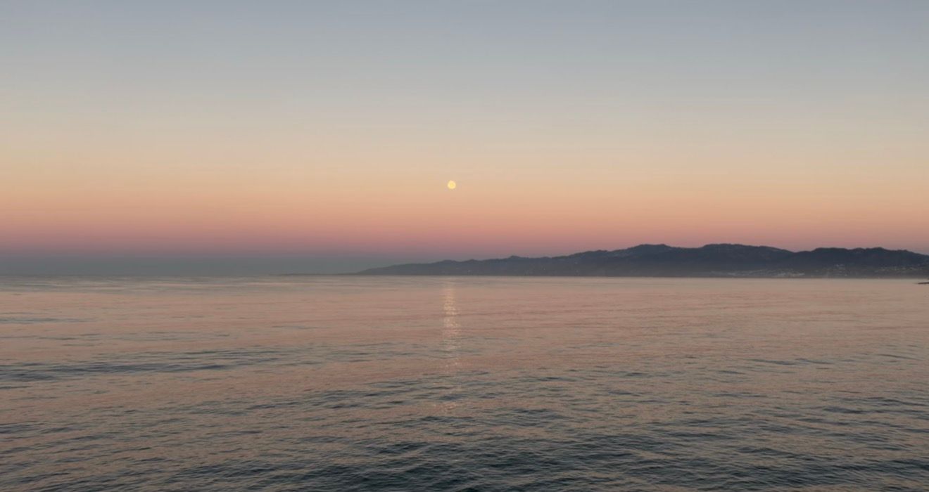 Full moon rising over calm ocean waters at sunset, with a pink and orange horizon glow and distant coastal mountains silhouetted.