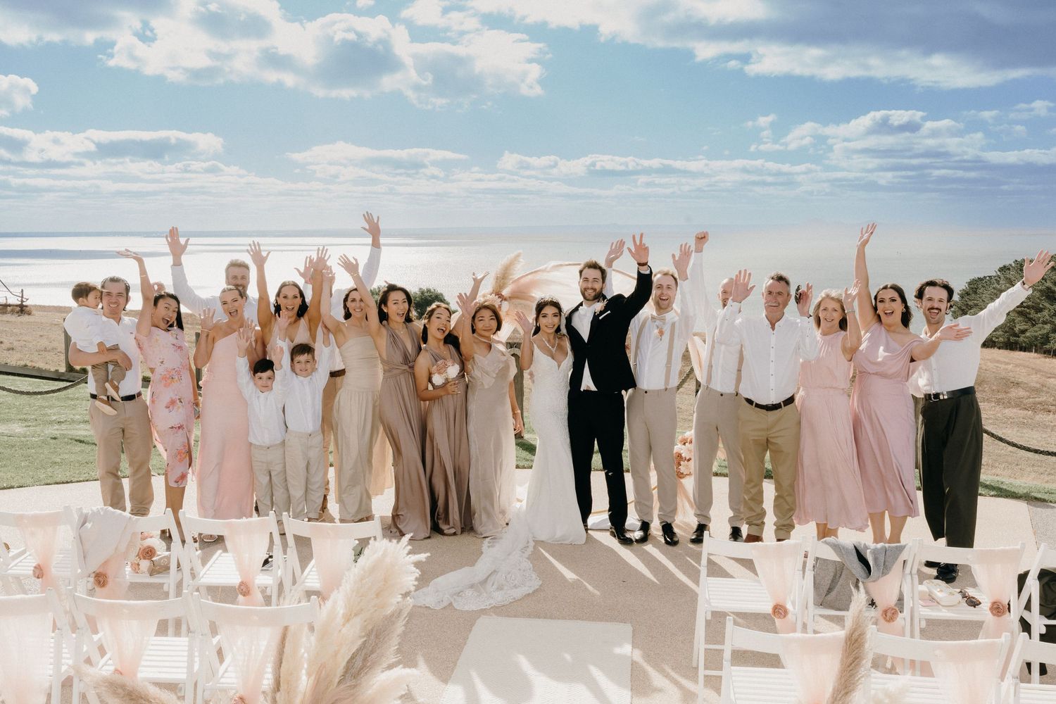 Wedding party celebrating outdoors with raised arms, bridesmaids in pink, ocean backdrop, white chairs, sunny day.