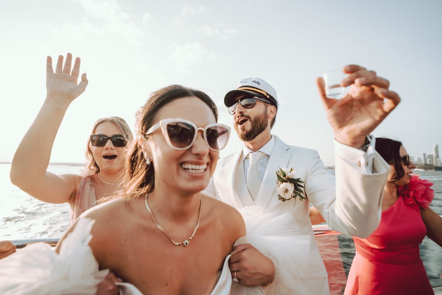 Joyful bride and groom celebrating on a boat with friends, bride wearing sunglasses and white dress.