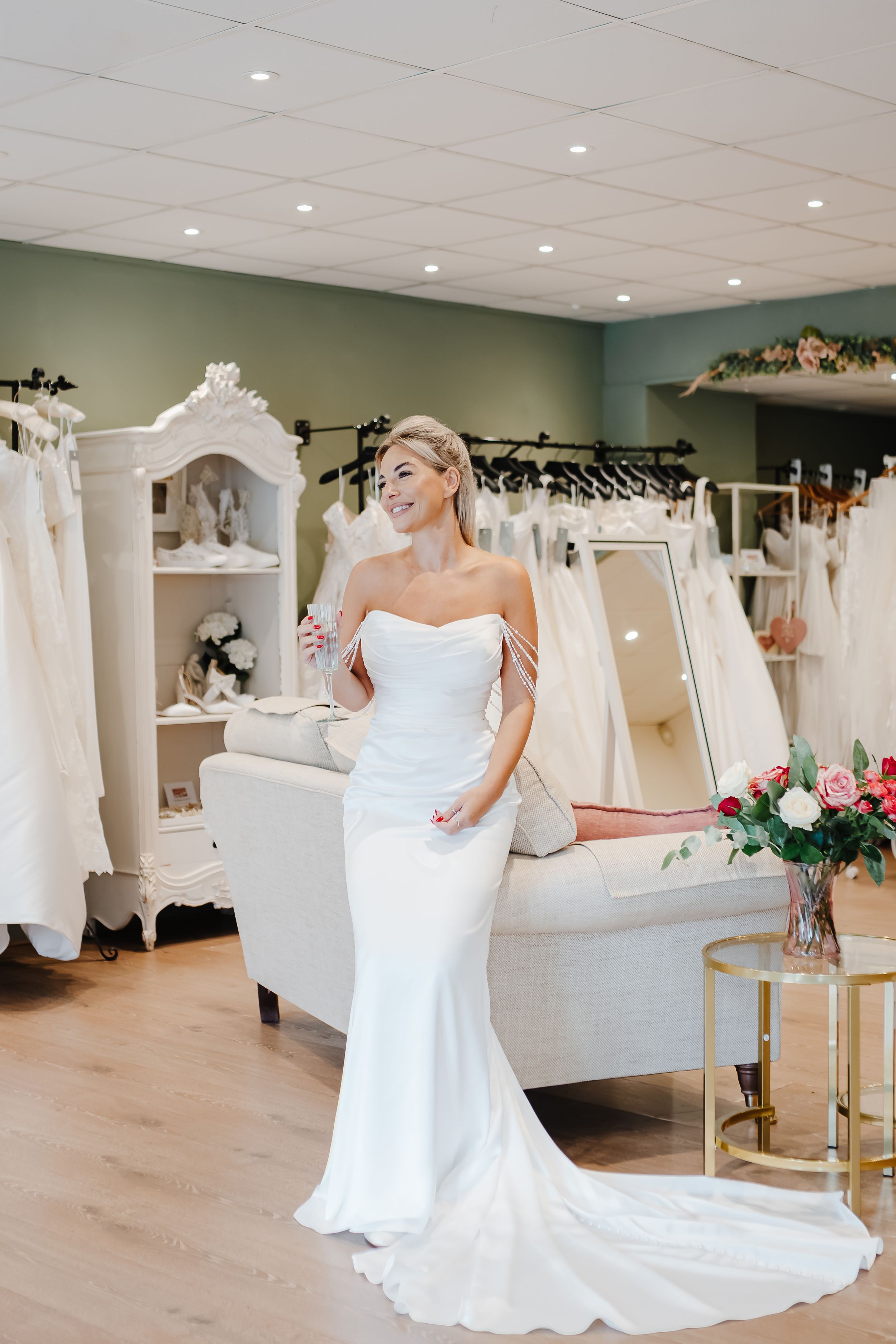 Bride wearing a sleek white off-shoulder mermaid wedding gown with a train, posing in an elegant bridal boutique.