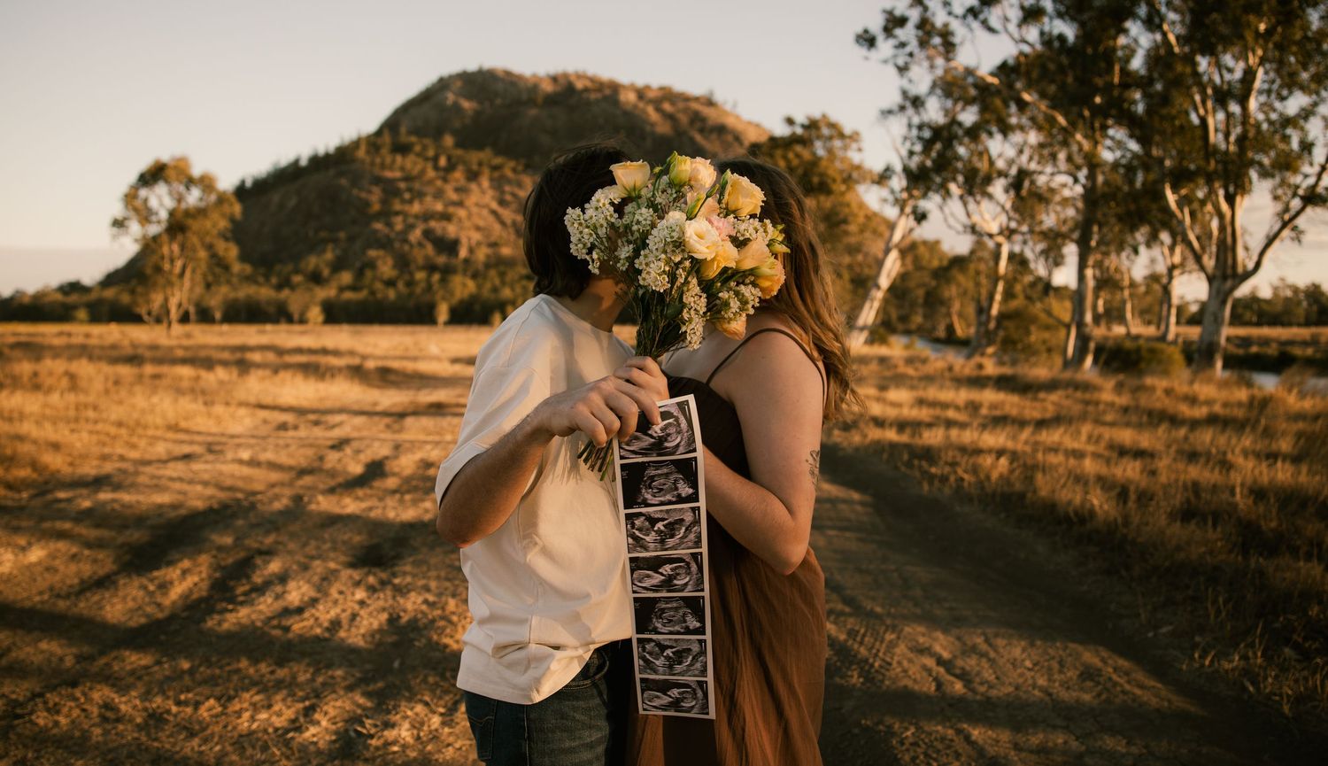 Couple kissing behind a bouquet of yellow flowers holding ultrasound photos in a golden field at sunset.