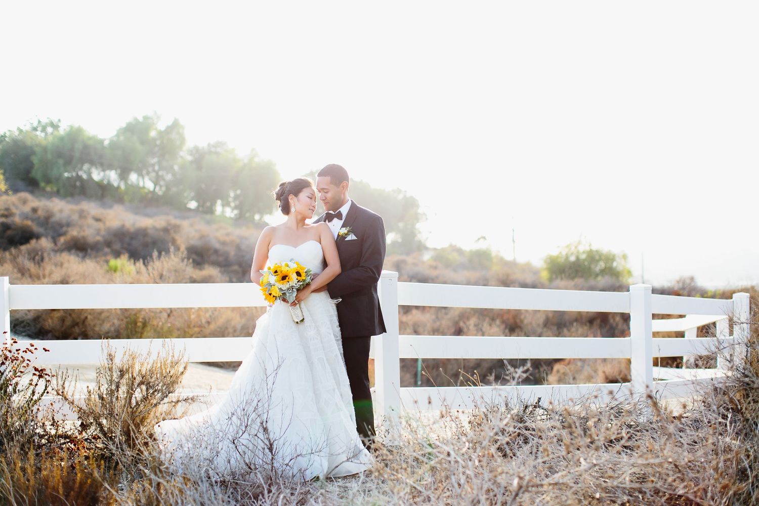 Wedding couple poses against rustic white fence during golden hour photography.
