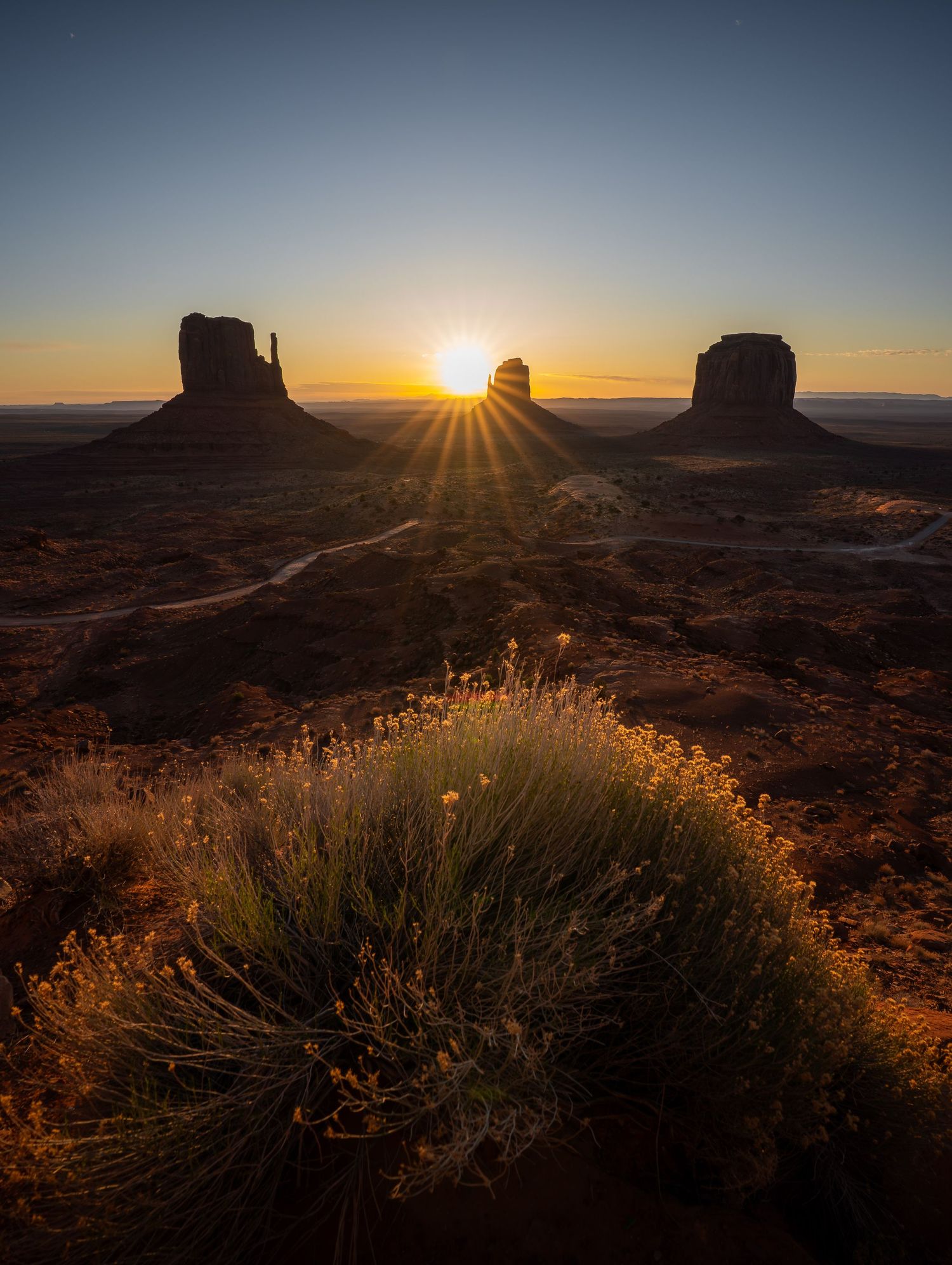 A series of silhouetted figures stand against a sunset sky viewed through natural rock arches in a desert landscape.