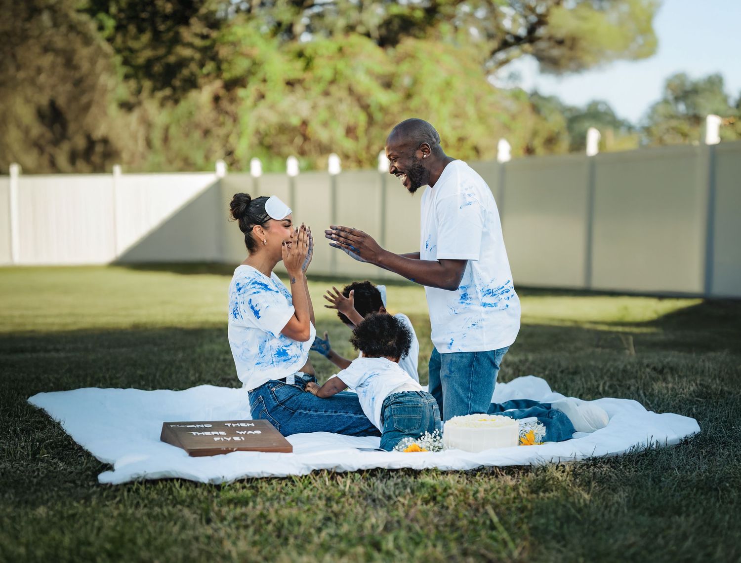 Happy family enjoying a picnic on a white blanket in a backyard garden, laughing and bonding together outdoors.