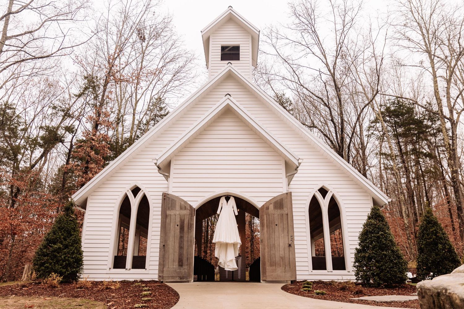 A white wedding dress hangs in the doorway of a charming white chapel with gothic arched windows in a wooded setting.