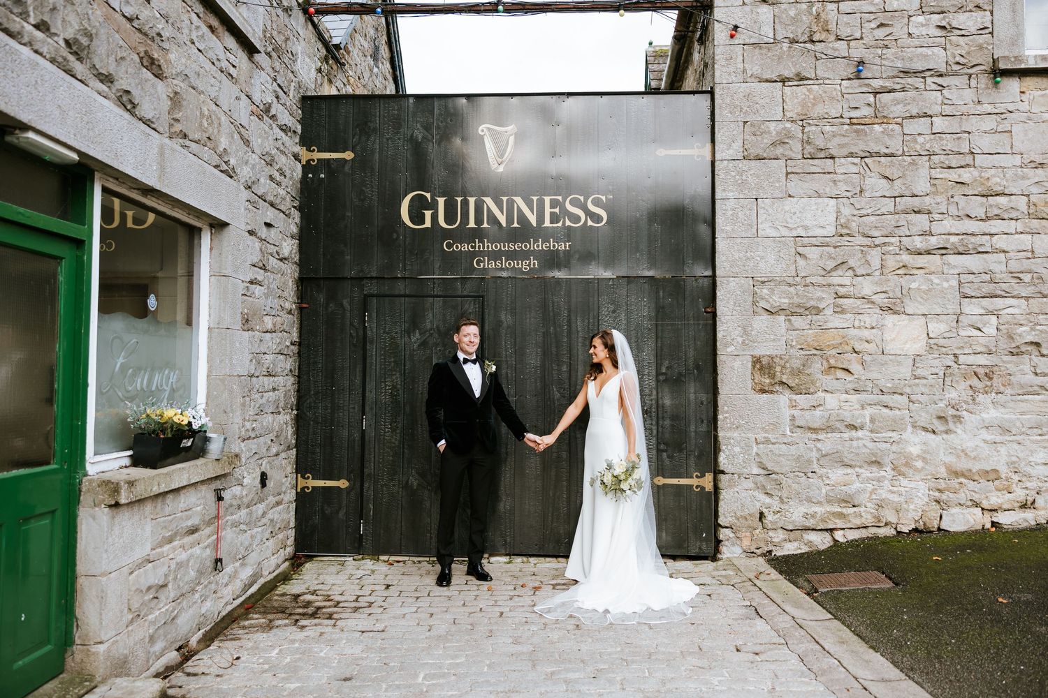 A wedding couple poses in front of rustic stone walls with green doors and a Guinness sign in an Irish setting. Castle Leslie