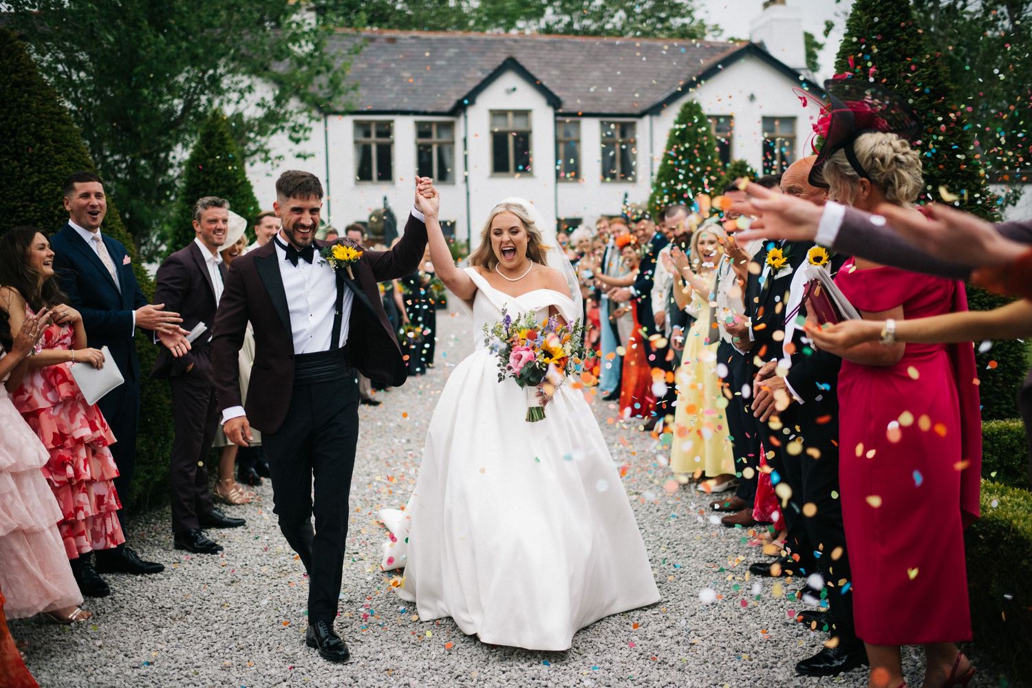 Wedding guests throw colorful confetti over the newlyweds during an outdoor celebration.