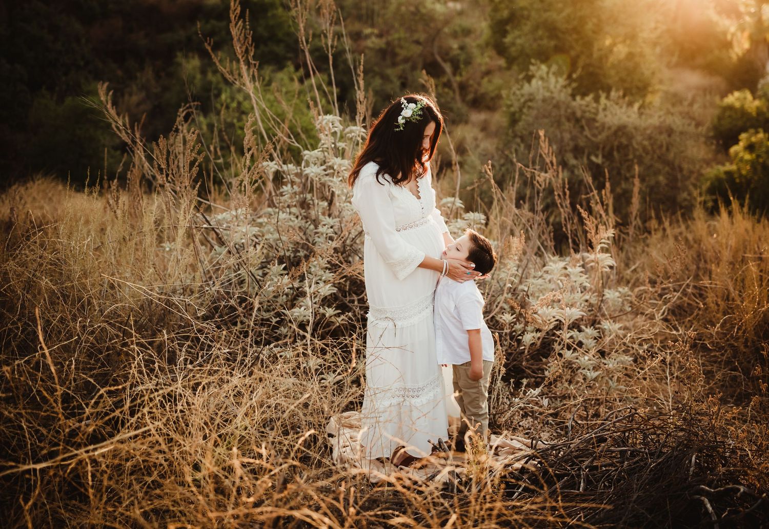 A couple in white outfits embraces during a golden hour photoshoot in a rustic outdoor setting with tall grass.