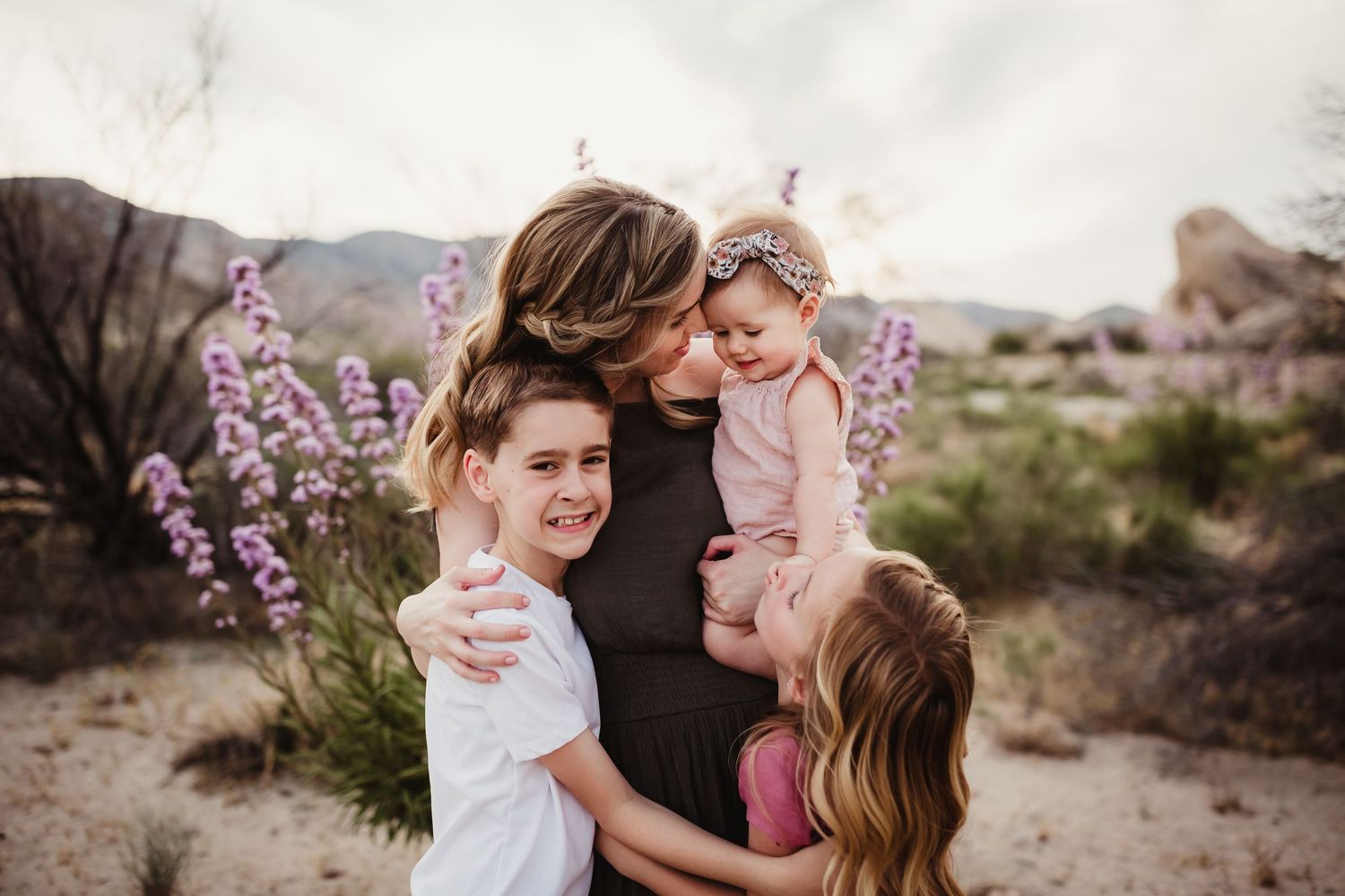 Mother and daughter share tender moments in a desert landscape with blooming purple wildflowers at sunset.