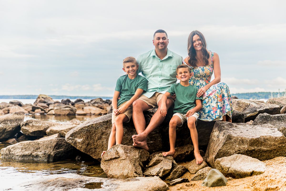 A family in coordinated beachwear sits together on coastal rocks with the ocean and cloudy sky in the background.