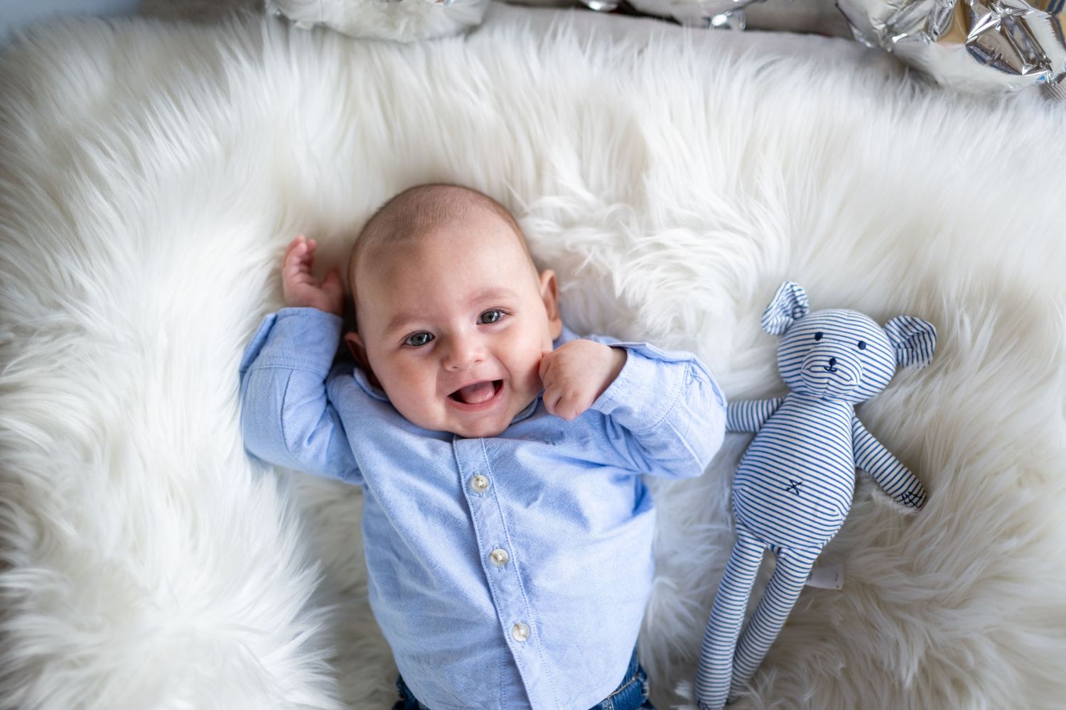Baby in light blue outfit lies on soft white fur blanket with plush toy teddy bear.