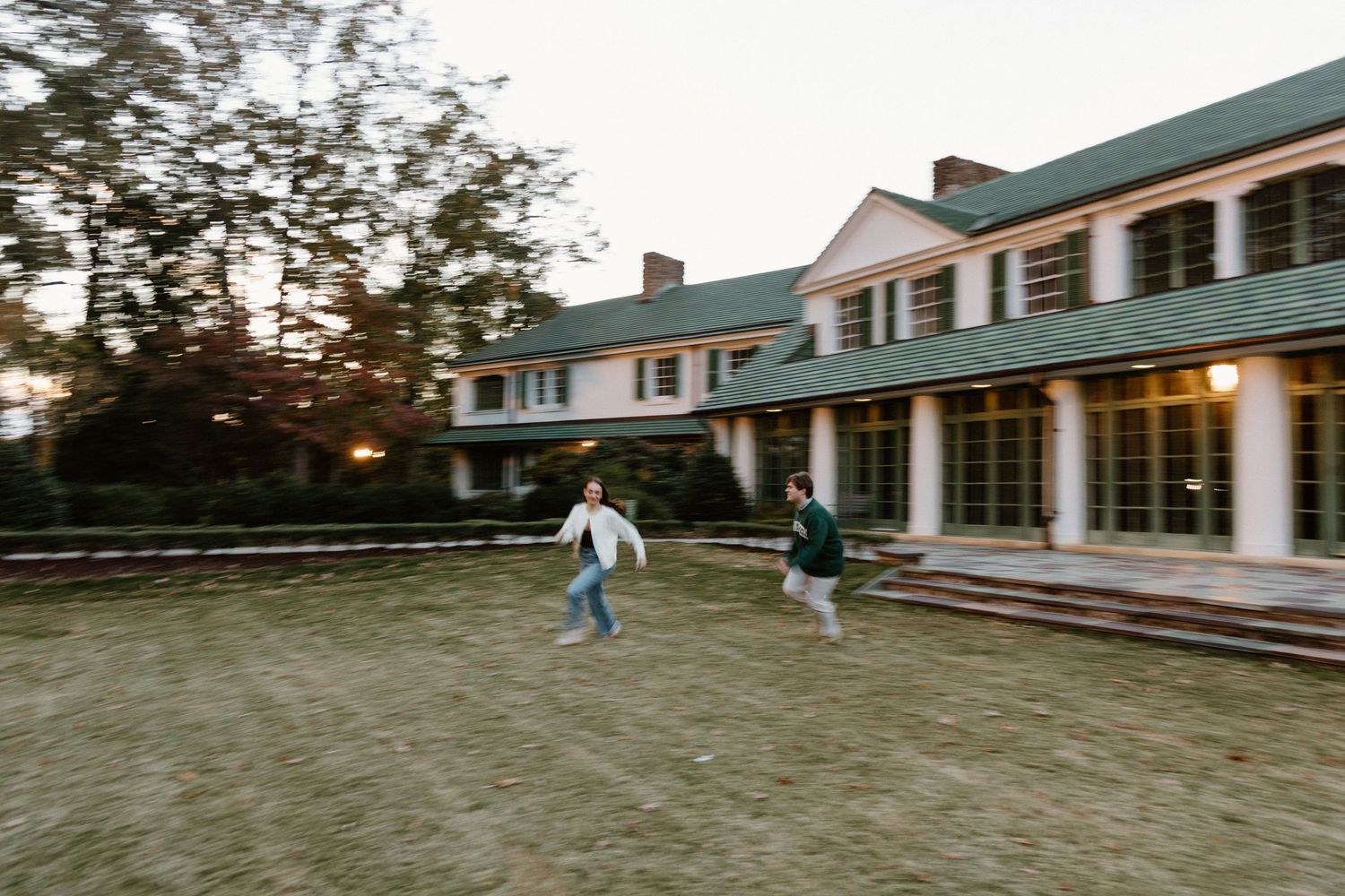 Two people run playfully across a lawn in front of a large white colonial-style building with green trim at dusk.